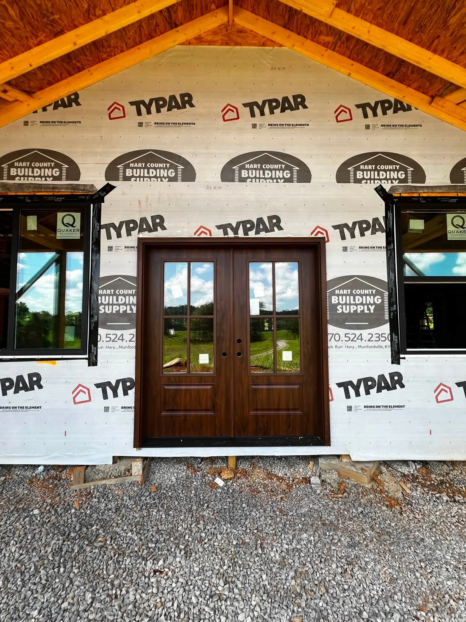 The exterior of a building under construction shows a wall with Typar house wrap and Hart County Building Supply logos, a wooden door frame, two windows, and a gravel ground.