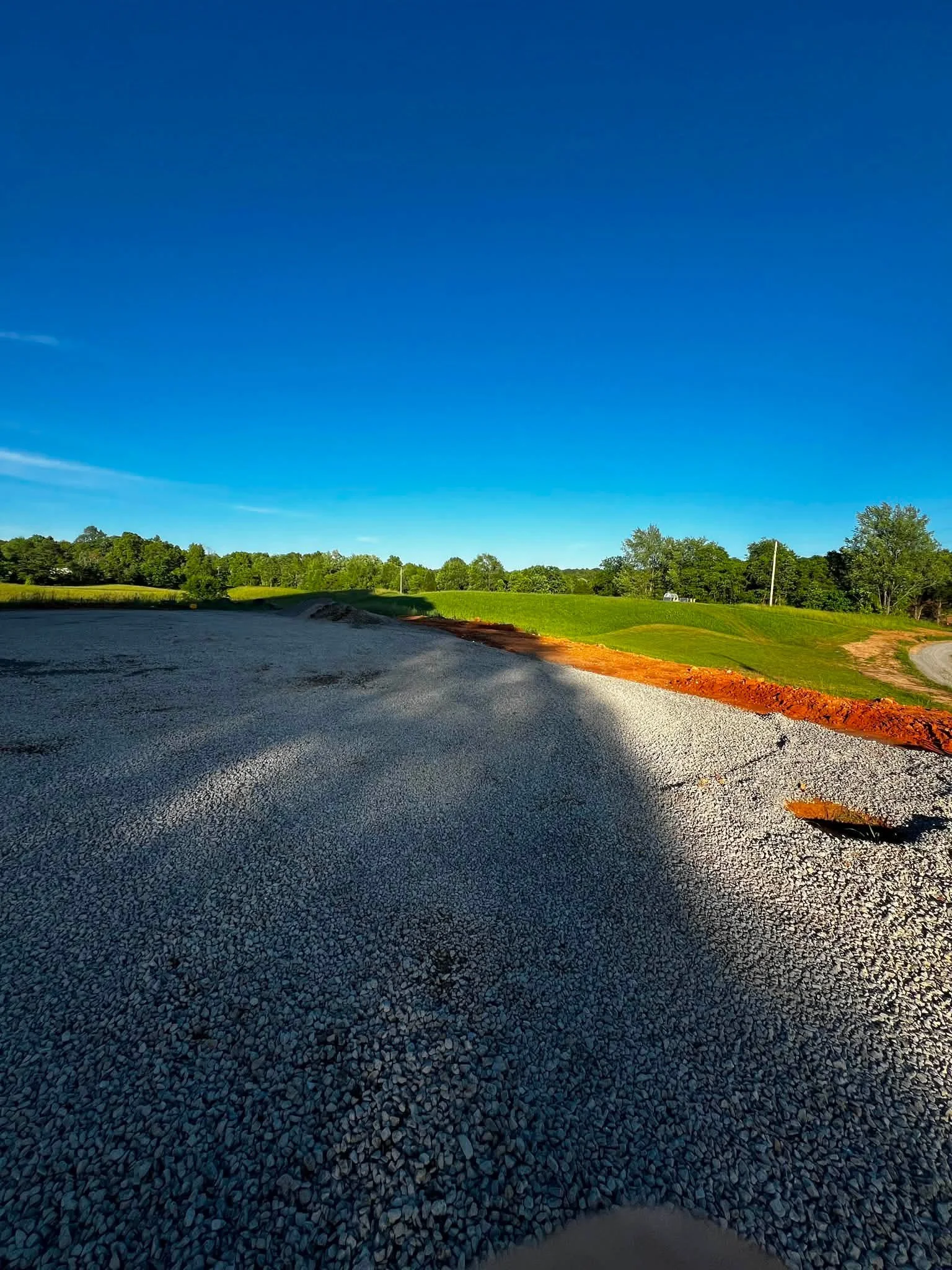 A gravel road curves through a green landscape with trees and grass, under a clear blue sky with no clouds.
