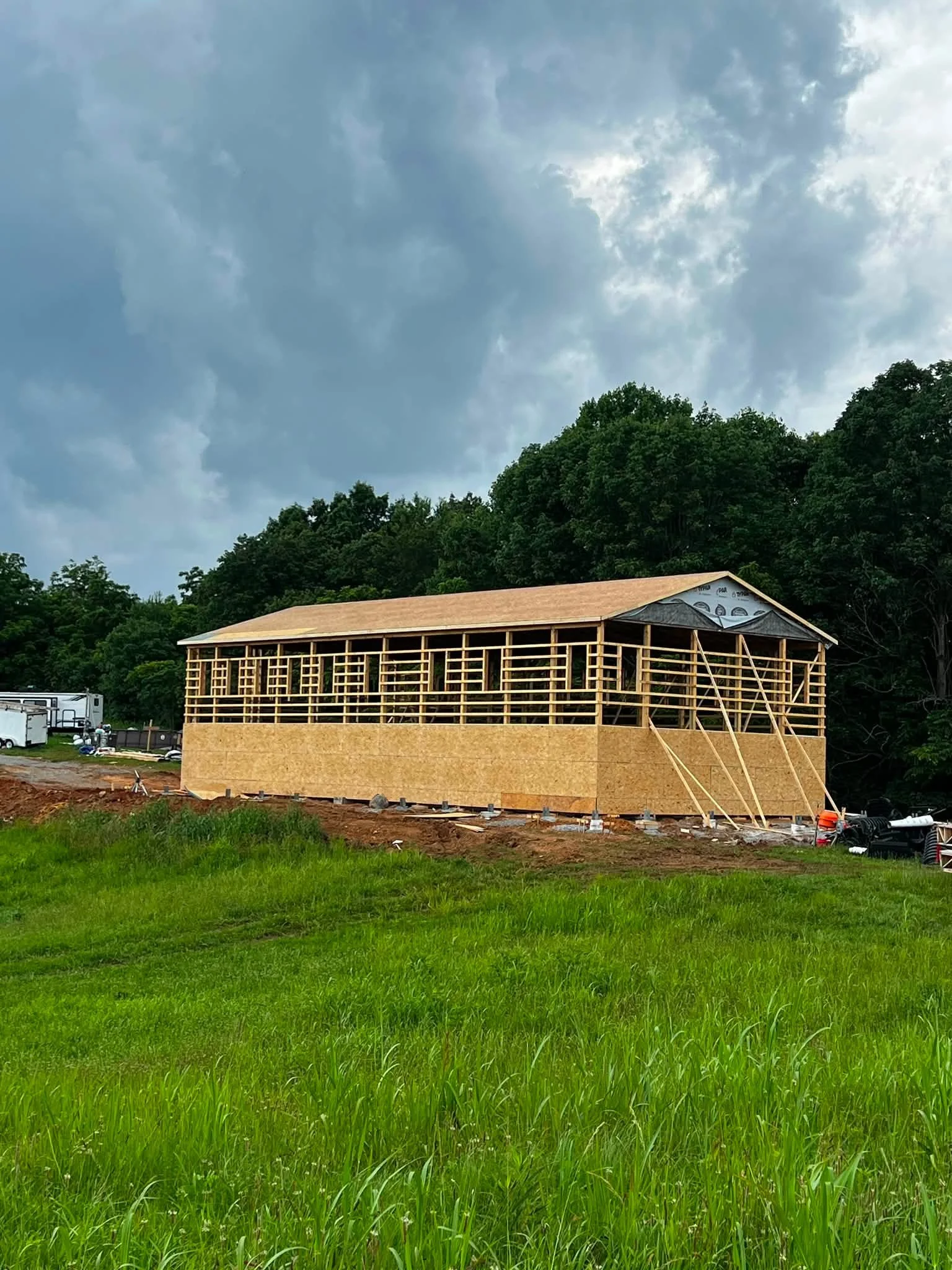 A house under construction with a wooden frame, surrounded by green grass and trees, with a cloudy sky overhead.