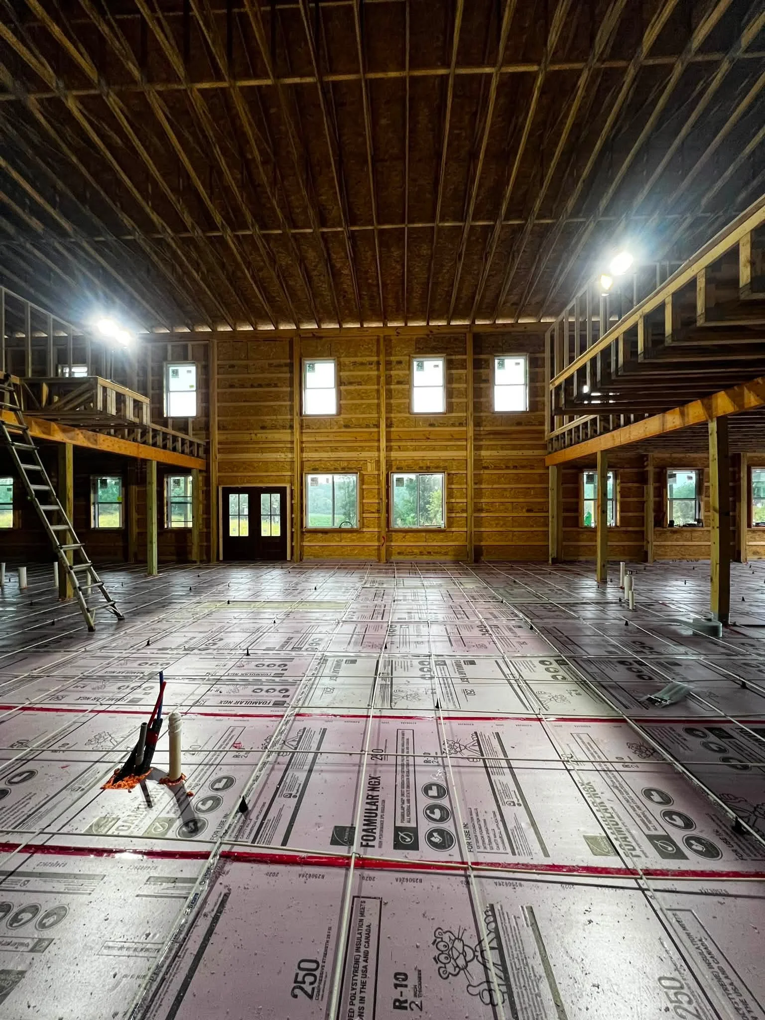 Interior of a building under construction with unfinished wooden framing and red piping, with a partially finished floor covered in construction plans or insulation sheathing.