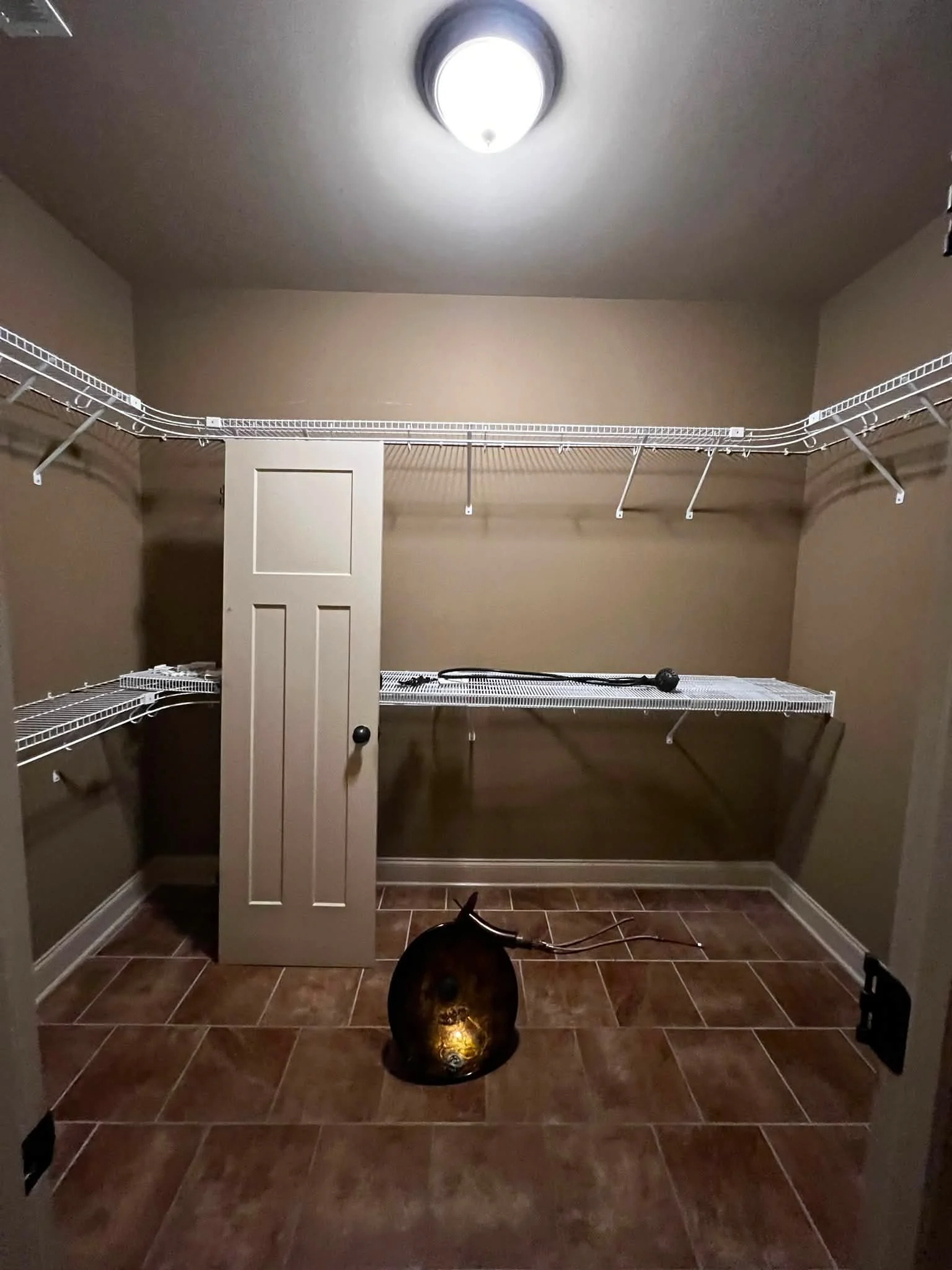 Empty walk-in closet with wire shelving, beige walls, and tiled floor, illuminated by ceiling light.