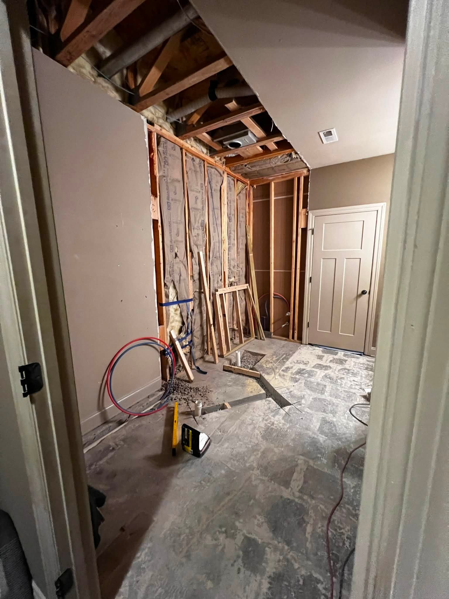 A room under construction with exposed wooden framing, pipes, and partially installed drywall. Tools and construction materials are visible on the floor.