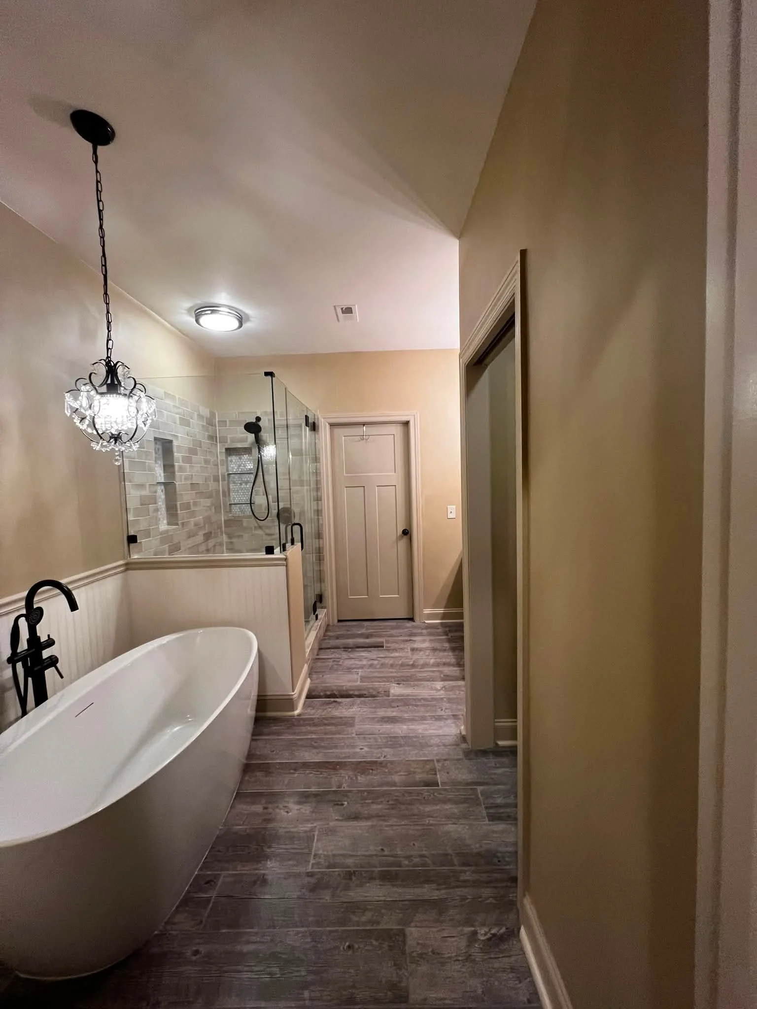 A bathroom with a clawfoot bathtub, a glass shower enclosure, beige walls, wood-like flooring, and a chandelier-style light fixture.