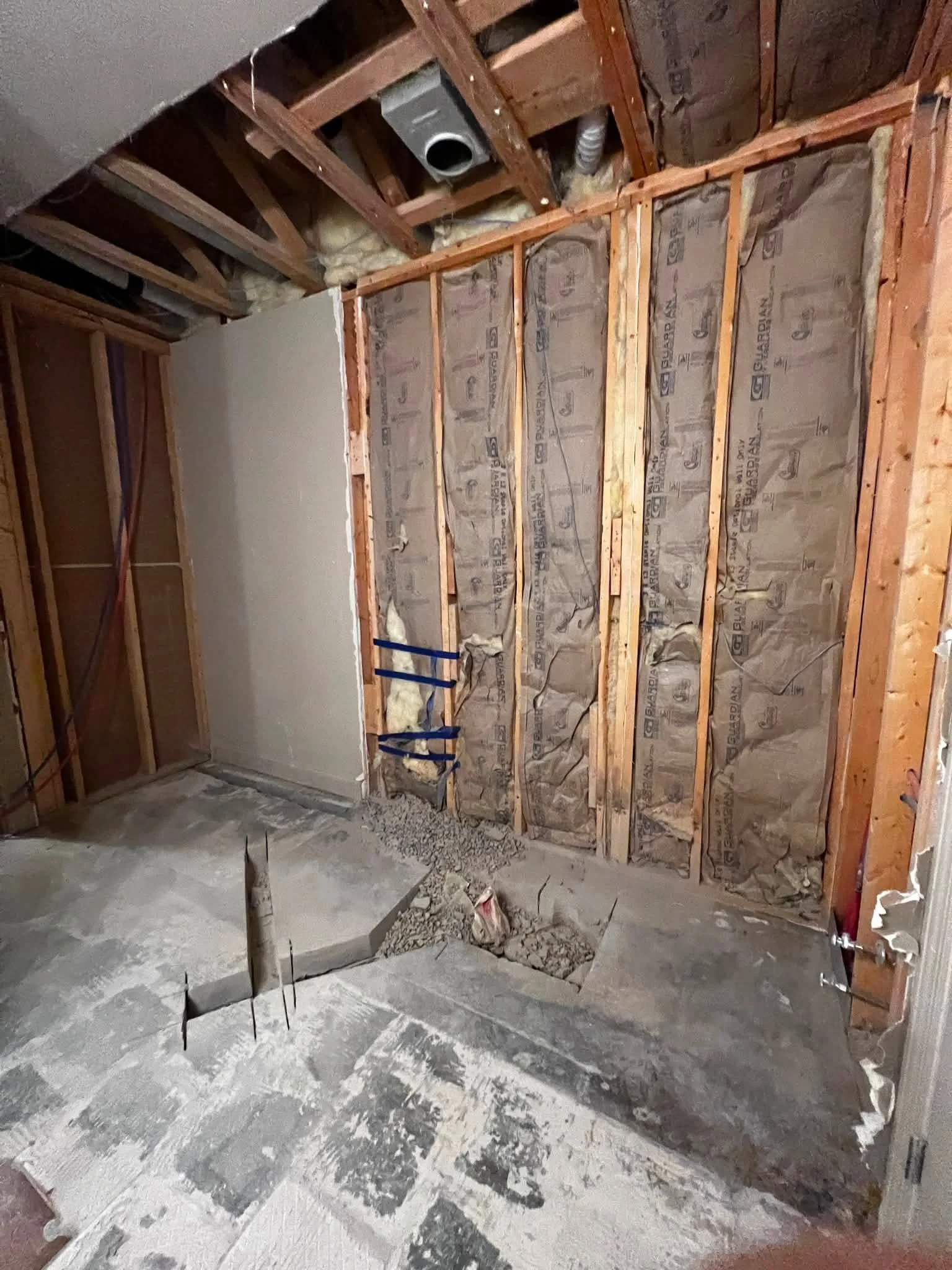 Interior of a building under construction or renovation with exposed wooden framing, insulation, and ductwork on the ceiling, and a concrete floor with debris and construction materials.