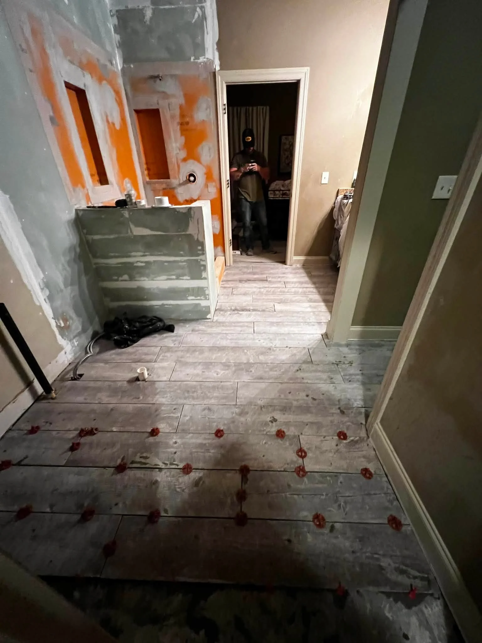 A room under renovation with partially painted walls and new flooring. There is a person in a dark hoodie and cap taking a photo via a mirror. The floor has red markers indicating future placements or measurements, and construction tools and material