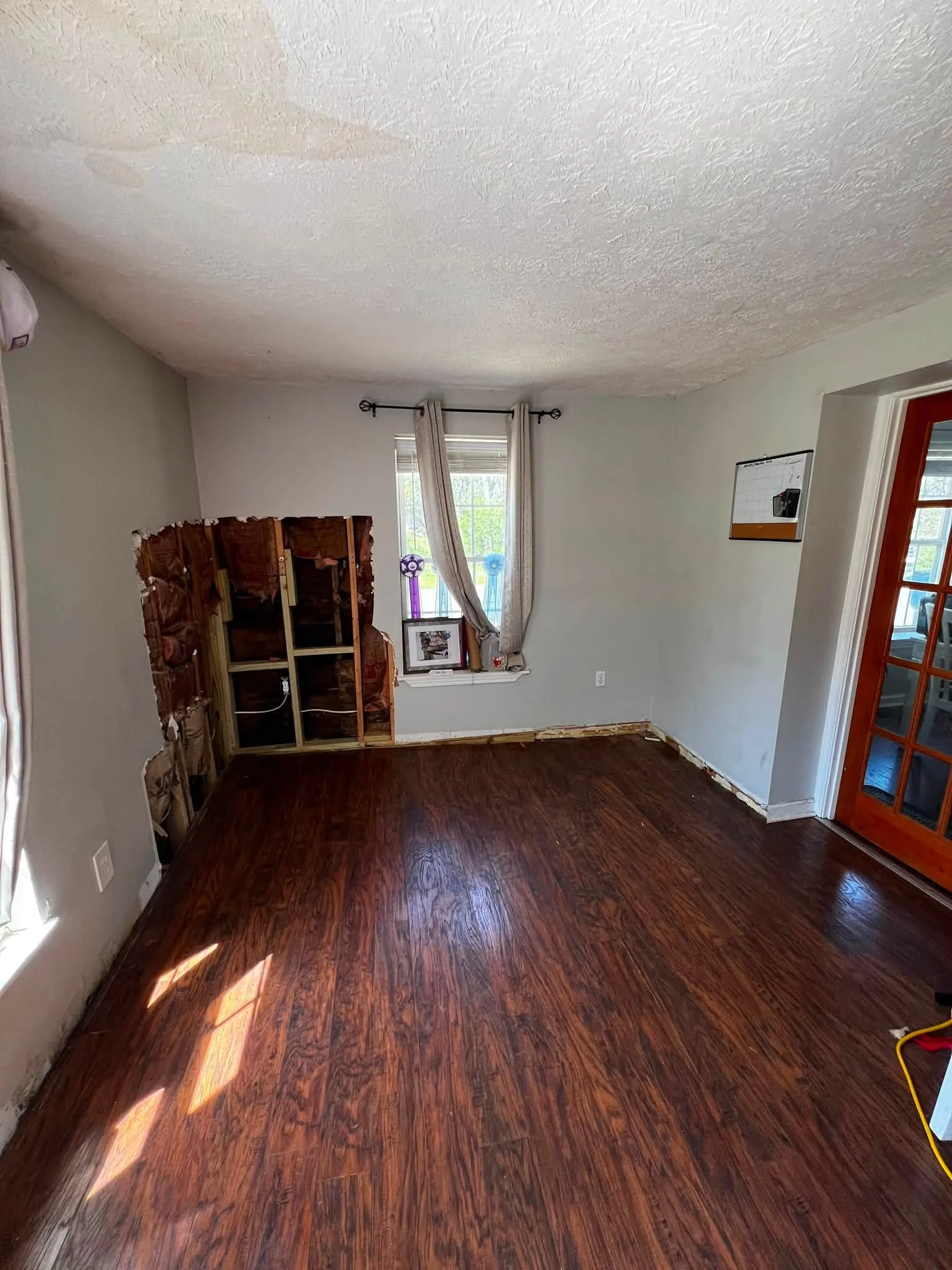 Room undergoing renovation with a partially removed wall, exposed studs, a window with curtains, hardwood flooring, and a glass-pane door.