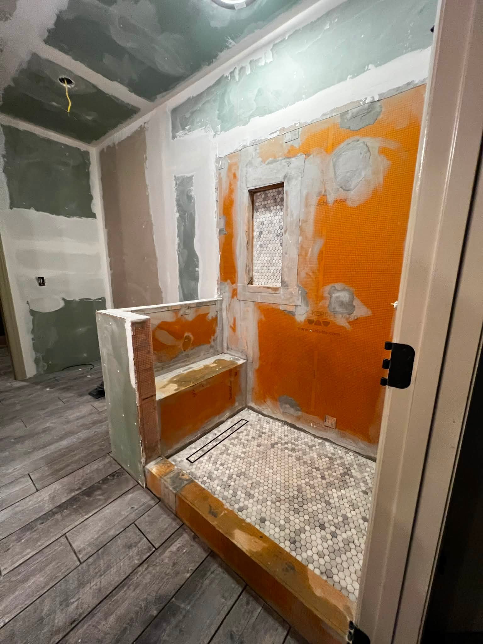 Bathroom under renovation with drywall installed and stairs leading to a shower area with white hexagonal tiles and orange waterproofing membrane, wood framing around the shower entrance, and gray wooden flooring.
