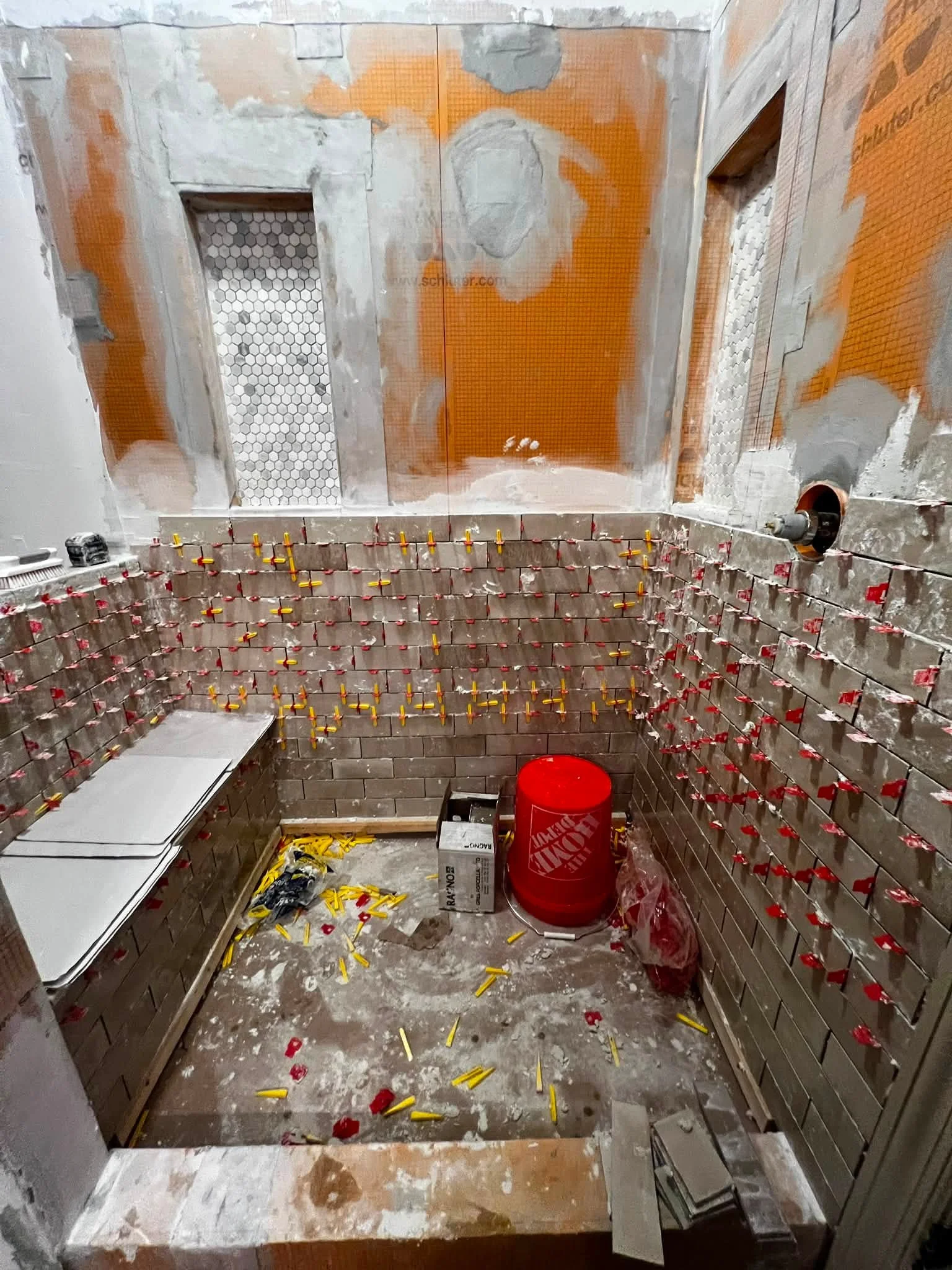 Under construction of a bathroom, with orange waterproofing membrane, partial wall and floor tiling, tools and construction materials scattered on the floor.