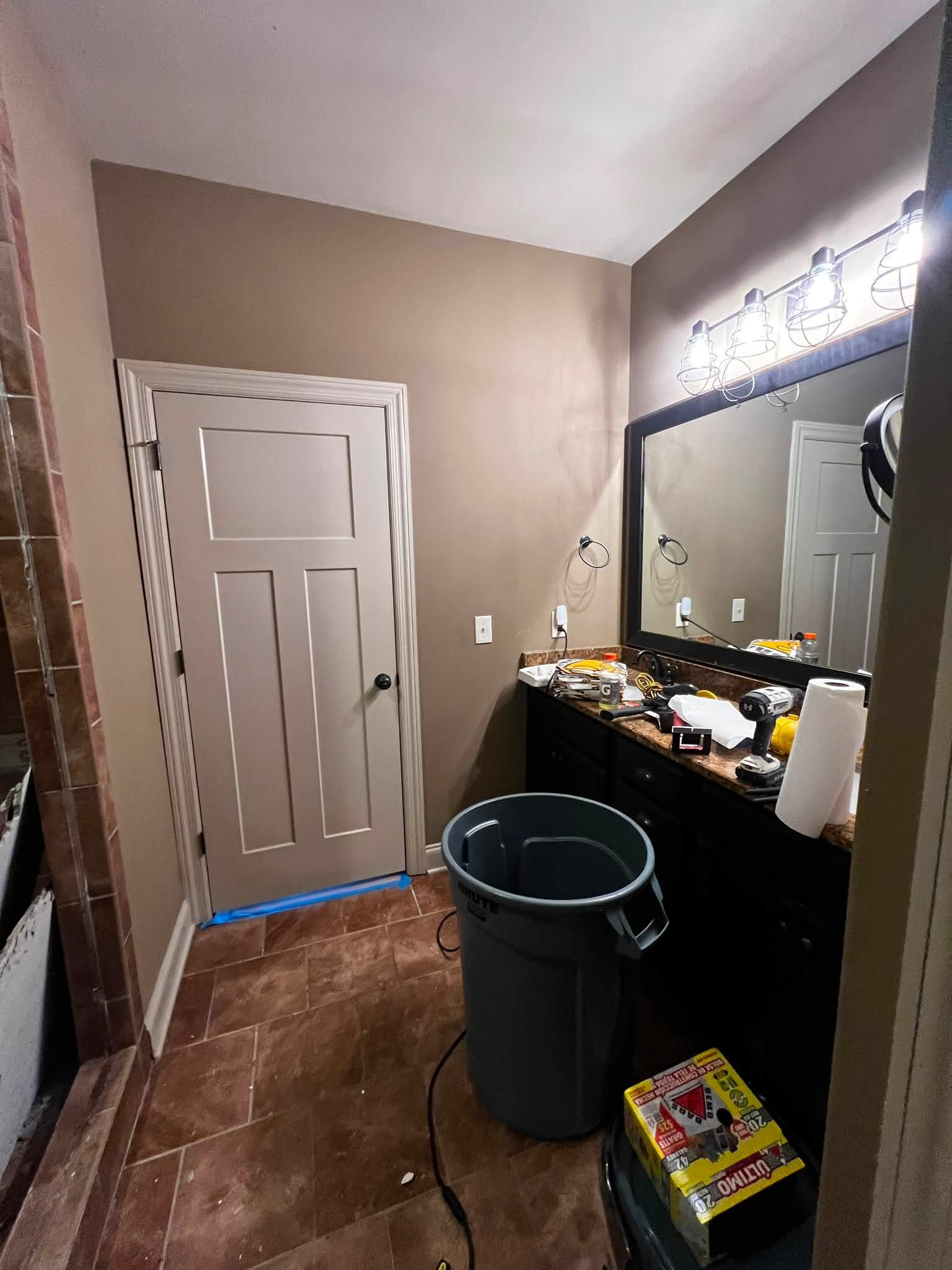 A bathroom vanity mirror with four vanity light fixtures above it, cluttered with various items including a power drill, a yellow box of tools, and a paper towel roll. A gray trash bin is in the foreground, and a closed white door is on the left side