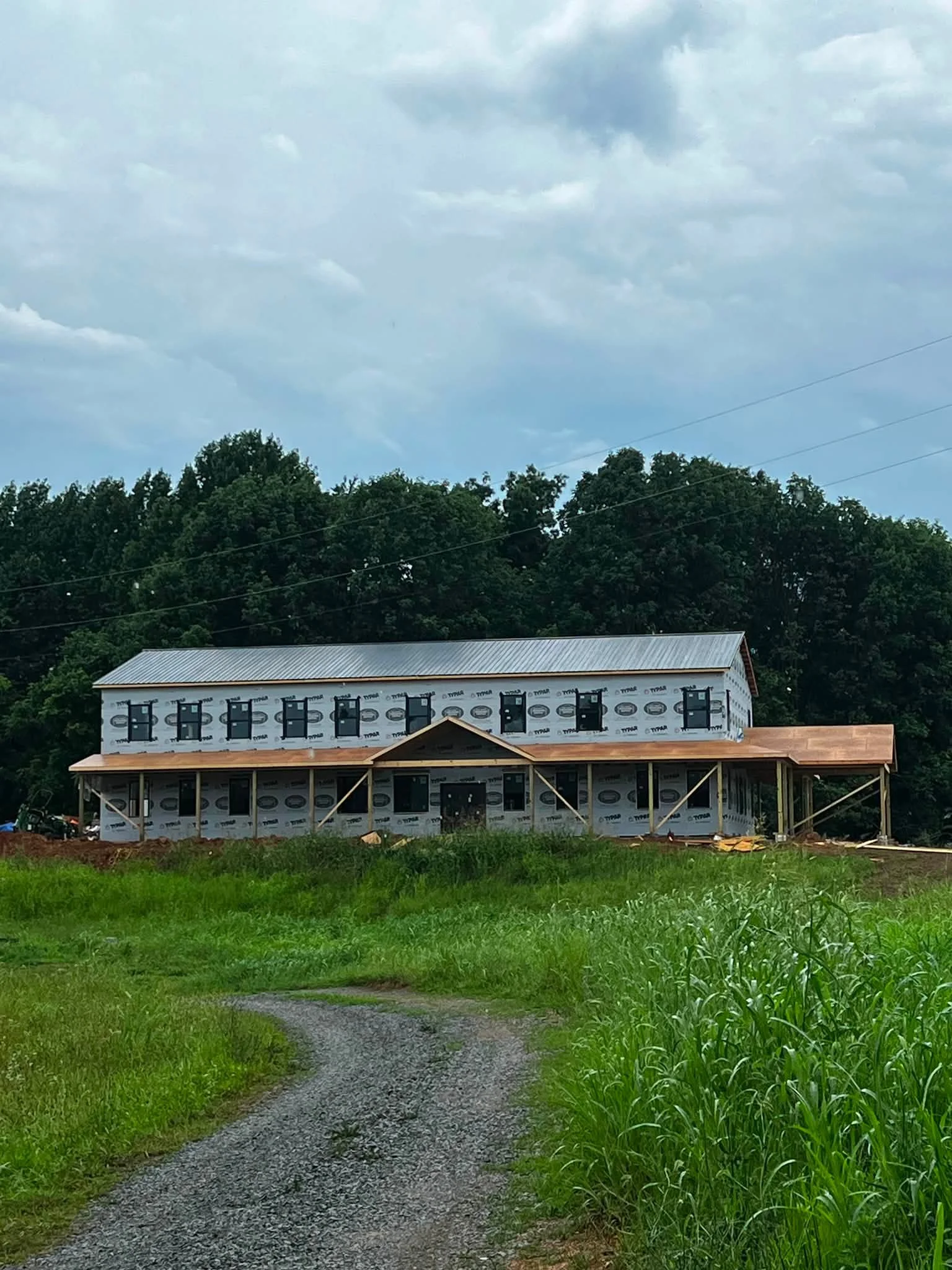 A house under construction with black window frames and a wrap-around porch, surrounded by green grass and trees, and a gravel driveway in the foreground under a cloudy sky.