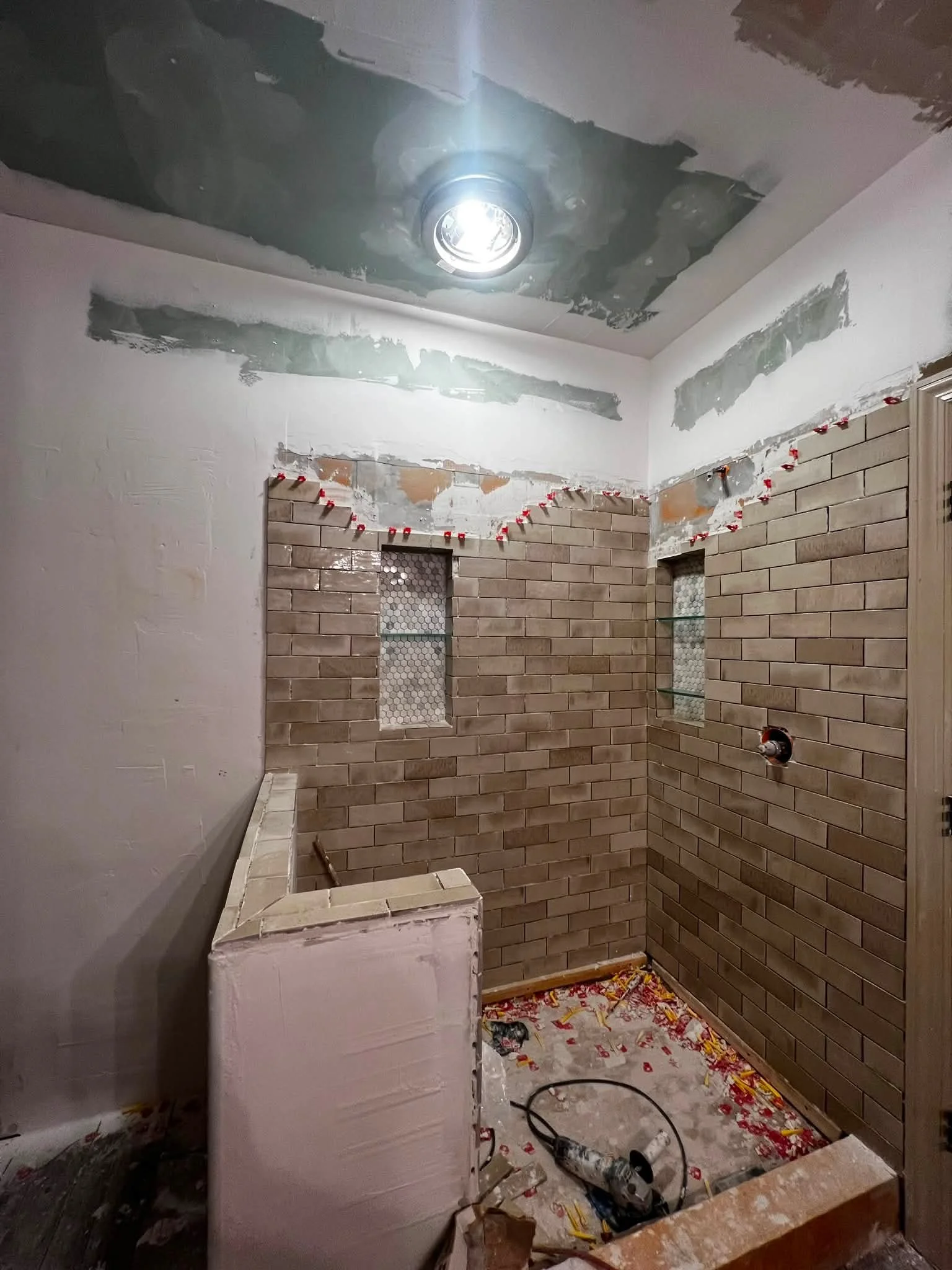 A bathroom under renovation with partially installed brick walls, construction tools, and debris on the floor. The ceiling has some incomplete drywall work with visible patches and a ceiling light fixture. The space is in the process of being finishe