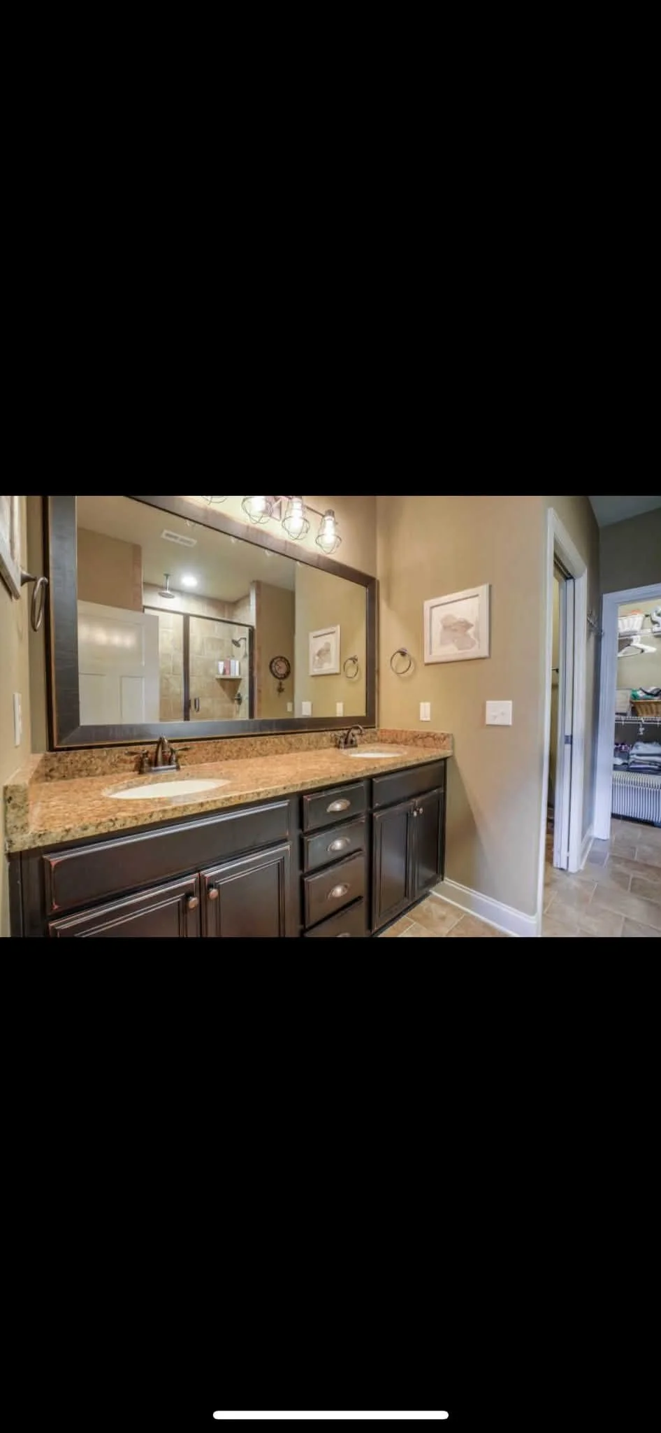 Bathroom with double vanity, granite countertop, large mirror, wall-mounted lights, dark wood cabinets, and adjacent walk-in closet.