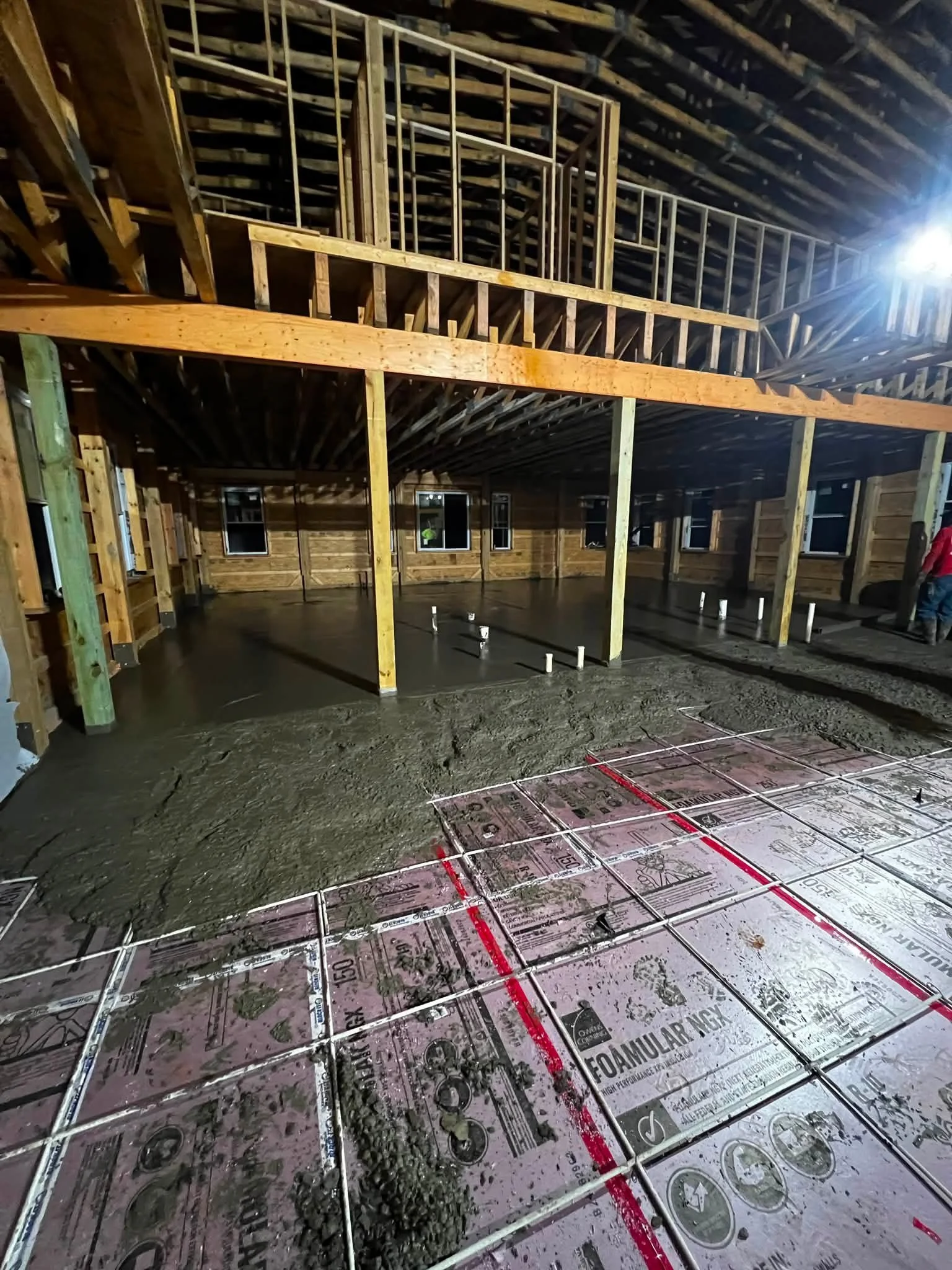 Construction site showing a concrete floor with heating or cooling pipes, wood framing for walls and an upper floor, with unfinished walls and windows, in a building under construction.