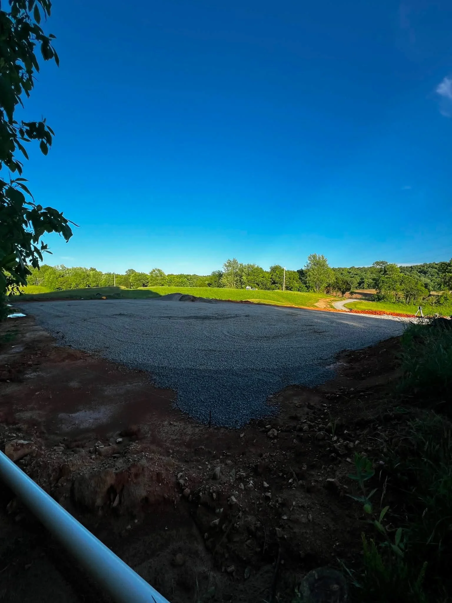 A gravel road under a clear blue sky with lush green trees in the distance, some tree branches and leaves partially frame the scene on the left side, and a blue pipe is visible at the bottom left corner.