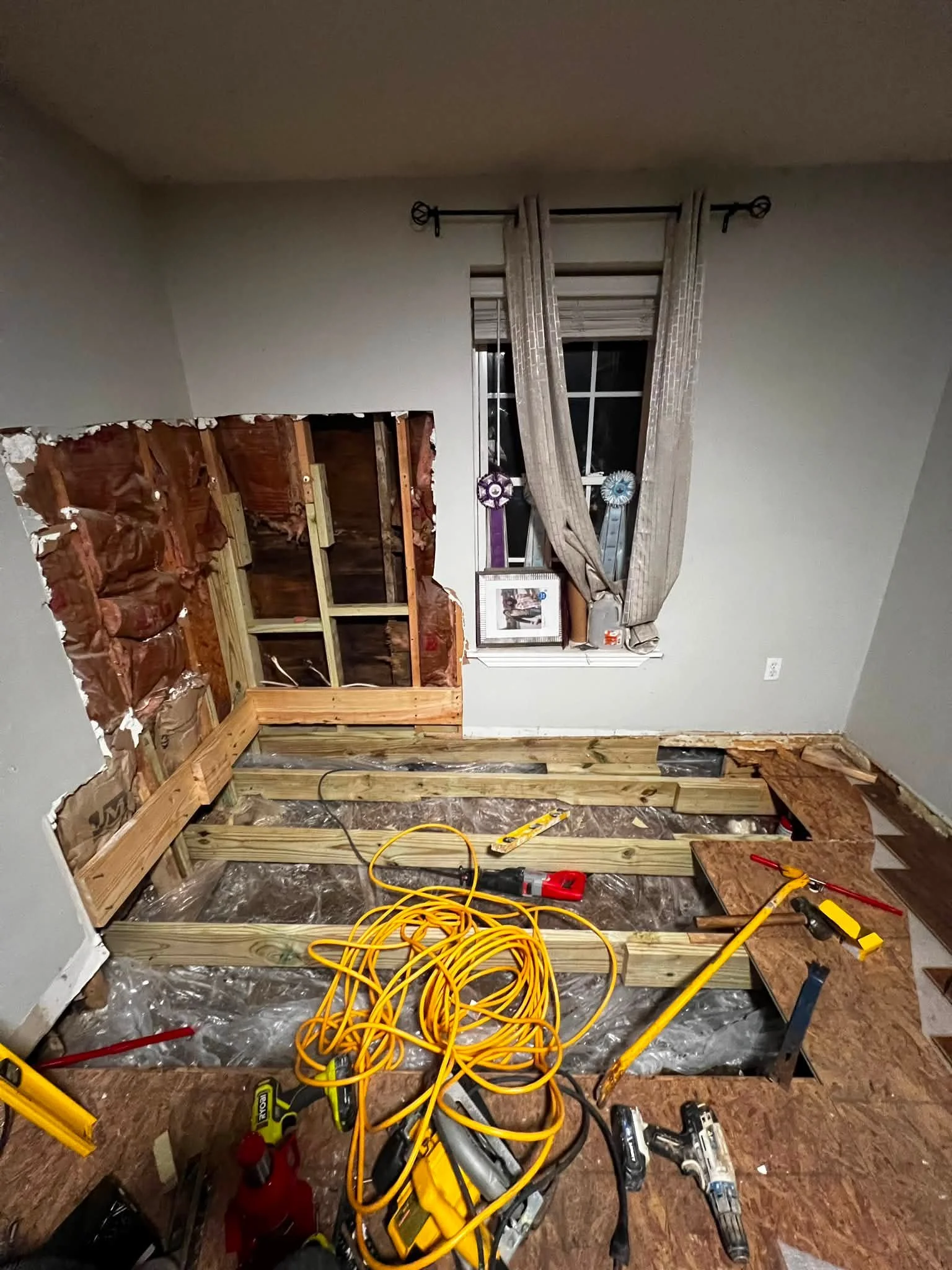 Room under construction with wooden framing, tools, and construction materials, including power drills, extension cords, and a partially torn wall covered with plastic