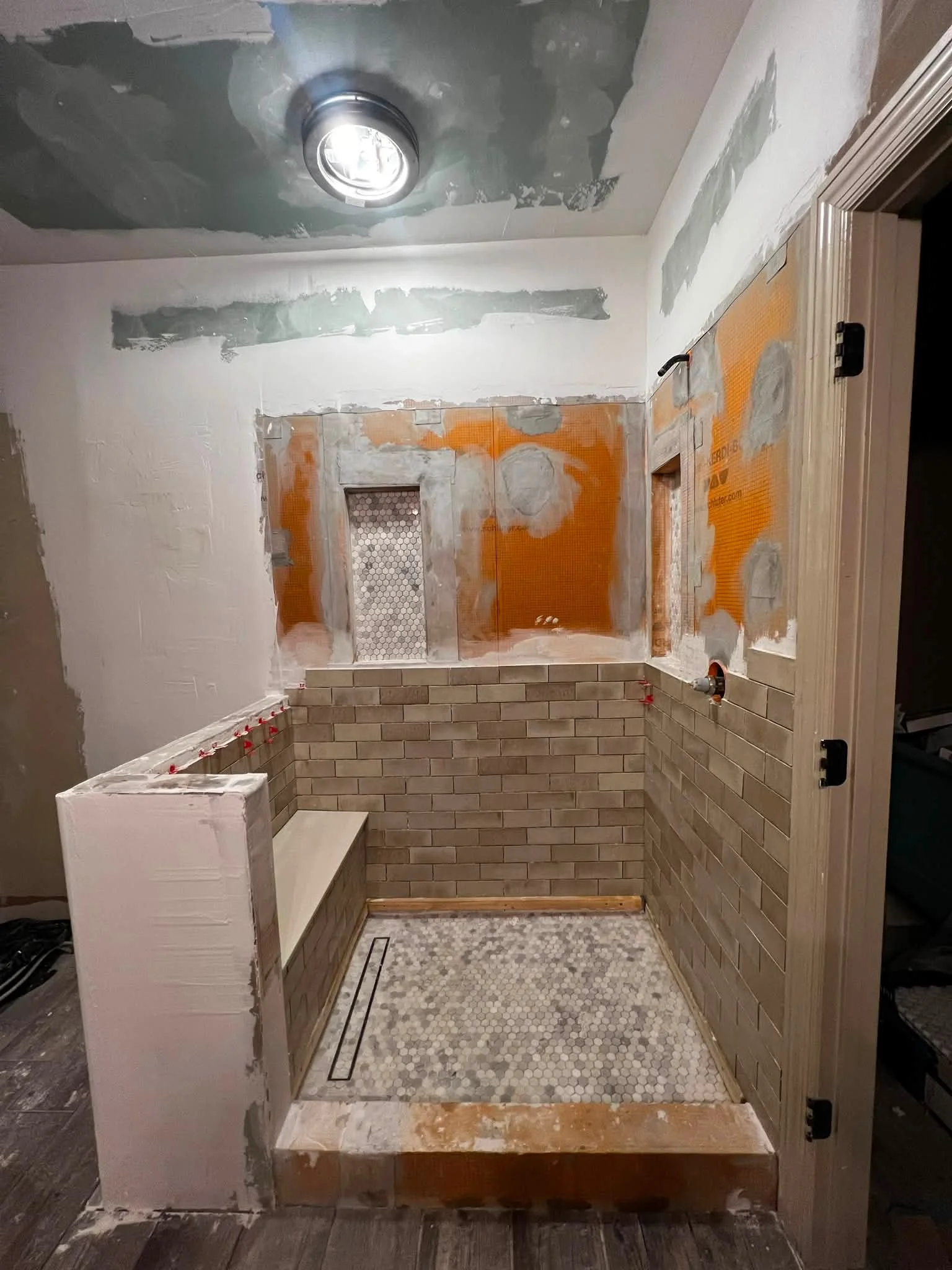 Bathroom under renovation with partially finished brick wall, small hexagonal tile floor, and drywall with patches of joint compound.