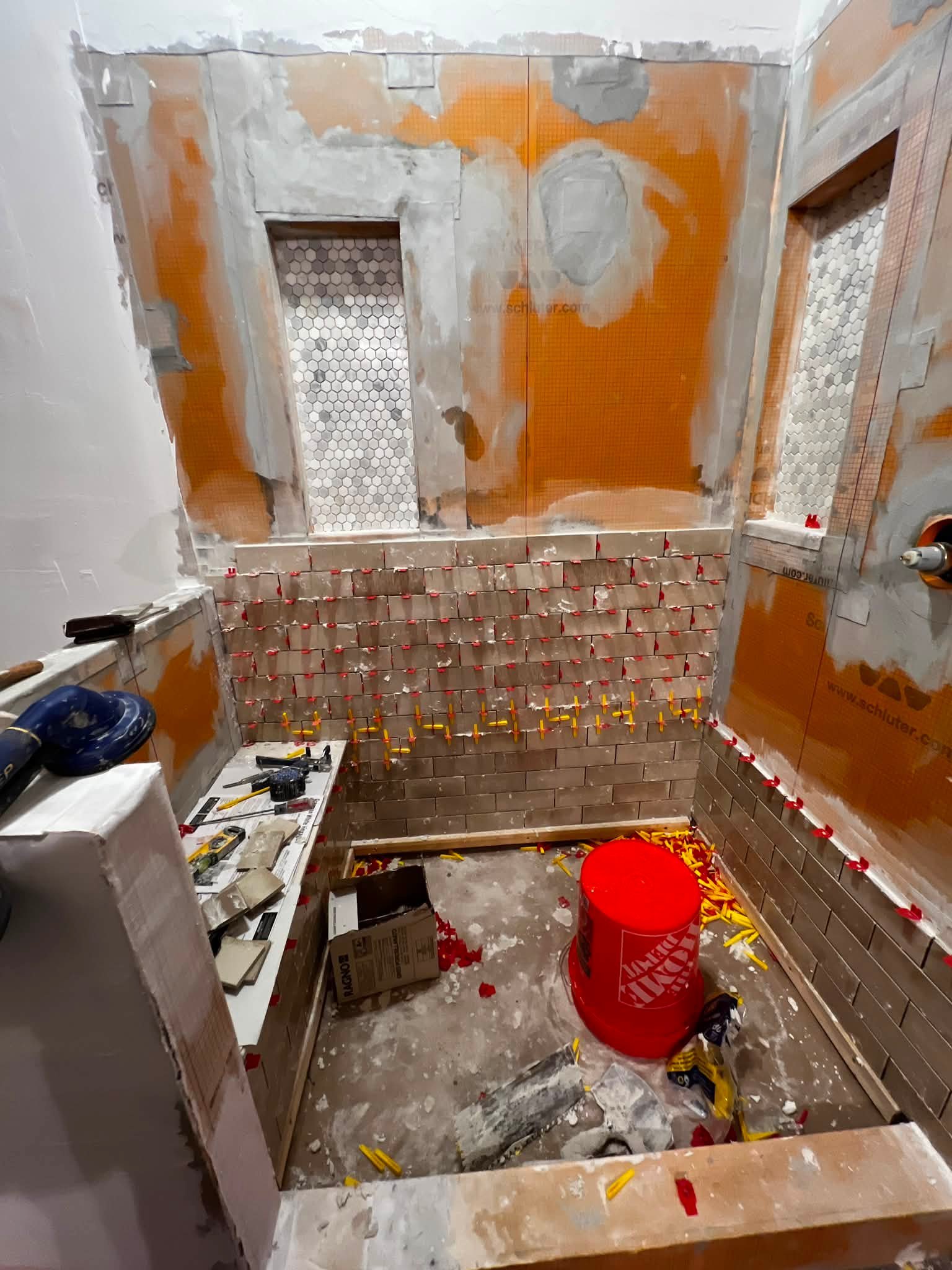 A room under construction with unfinished walls, tile installation in progress, tools, and construction materials scattered on the floor and countertops.