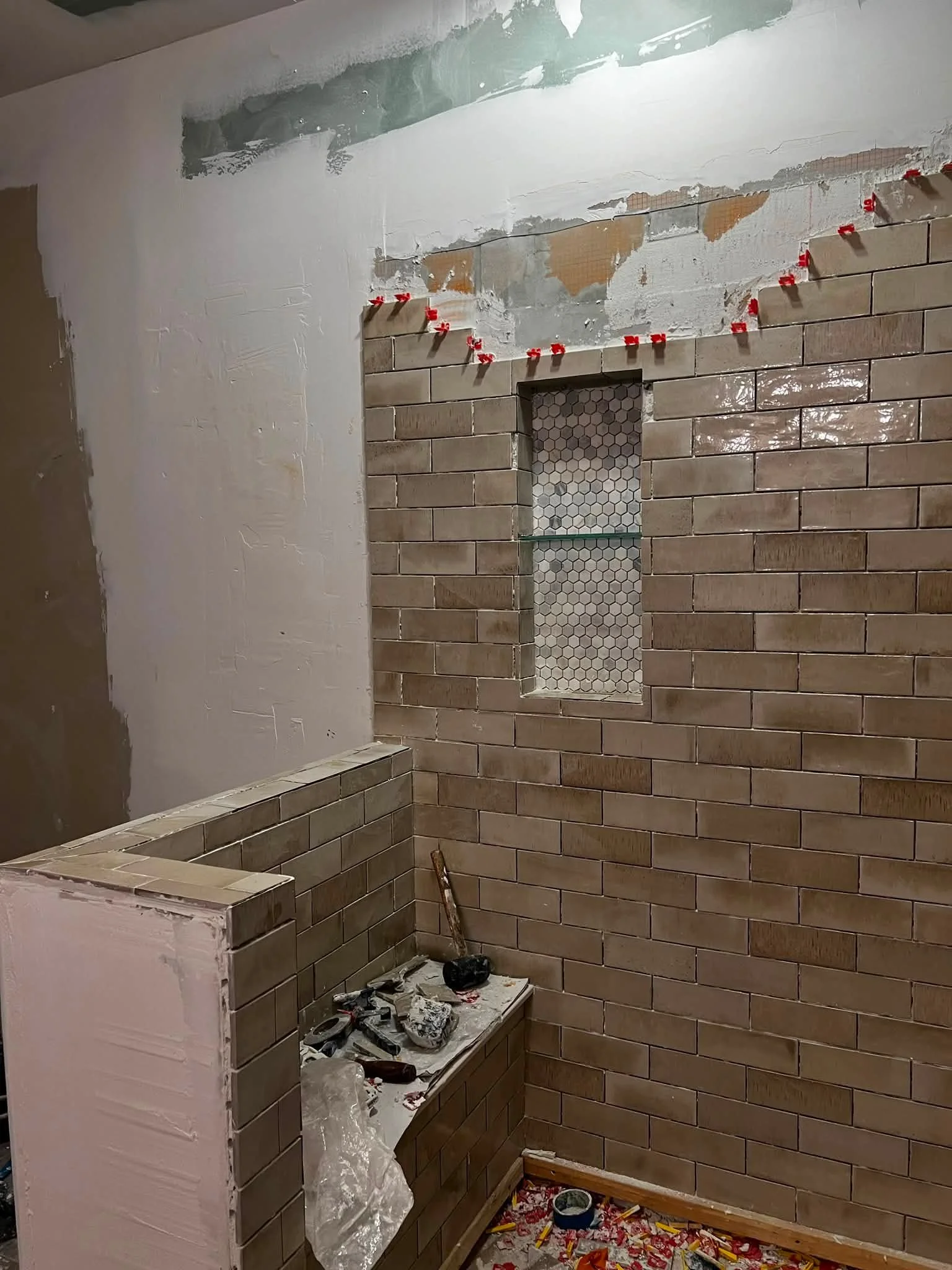 A wall under construction with beige brick tiles partially installed, a niche with hexagonal tiles, and a ledge with tools and construction materials in a room undergoing renovation.