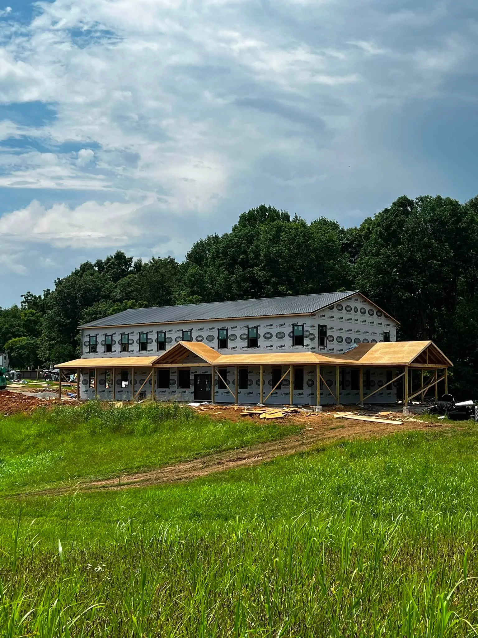 A house under construction with a metal roof and taped exterior walls, surrounded by green grass and trees, under a partly cloudy sky.