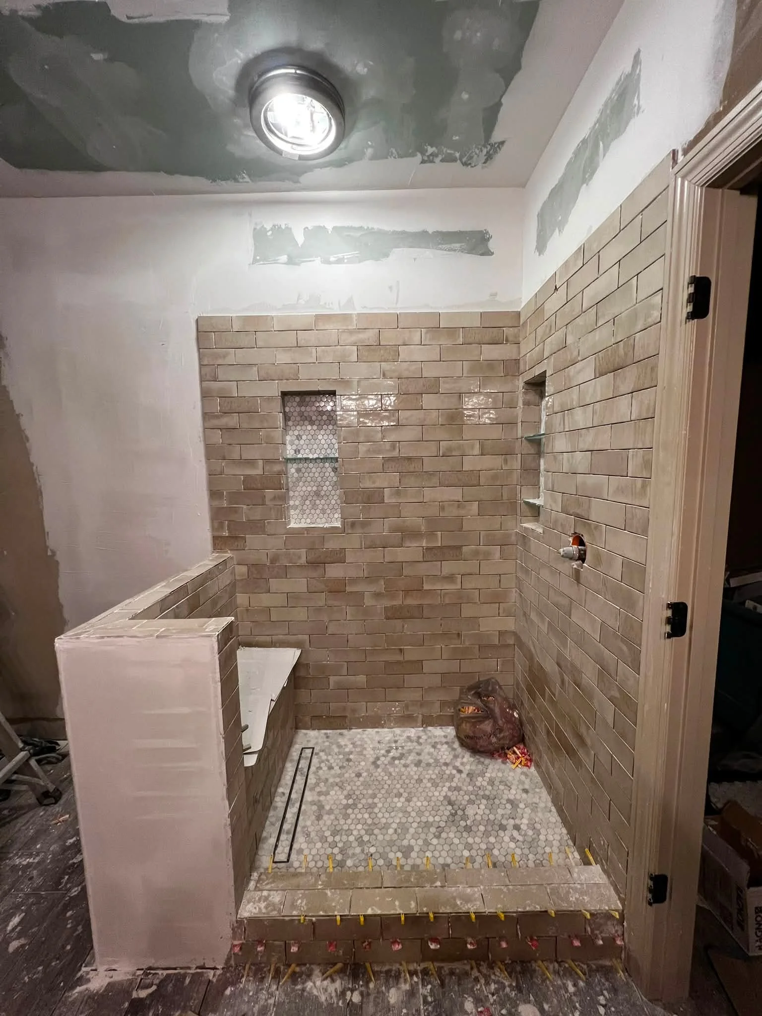 Bathroom under renovation with partially tiled wall, a small niche with hexagonal tiles, shower area with hex tiles on the floor, and construction materials on the floor.