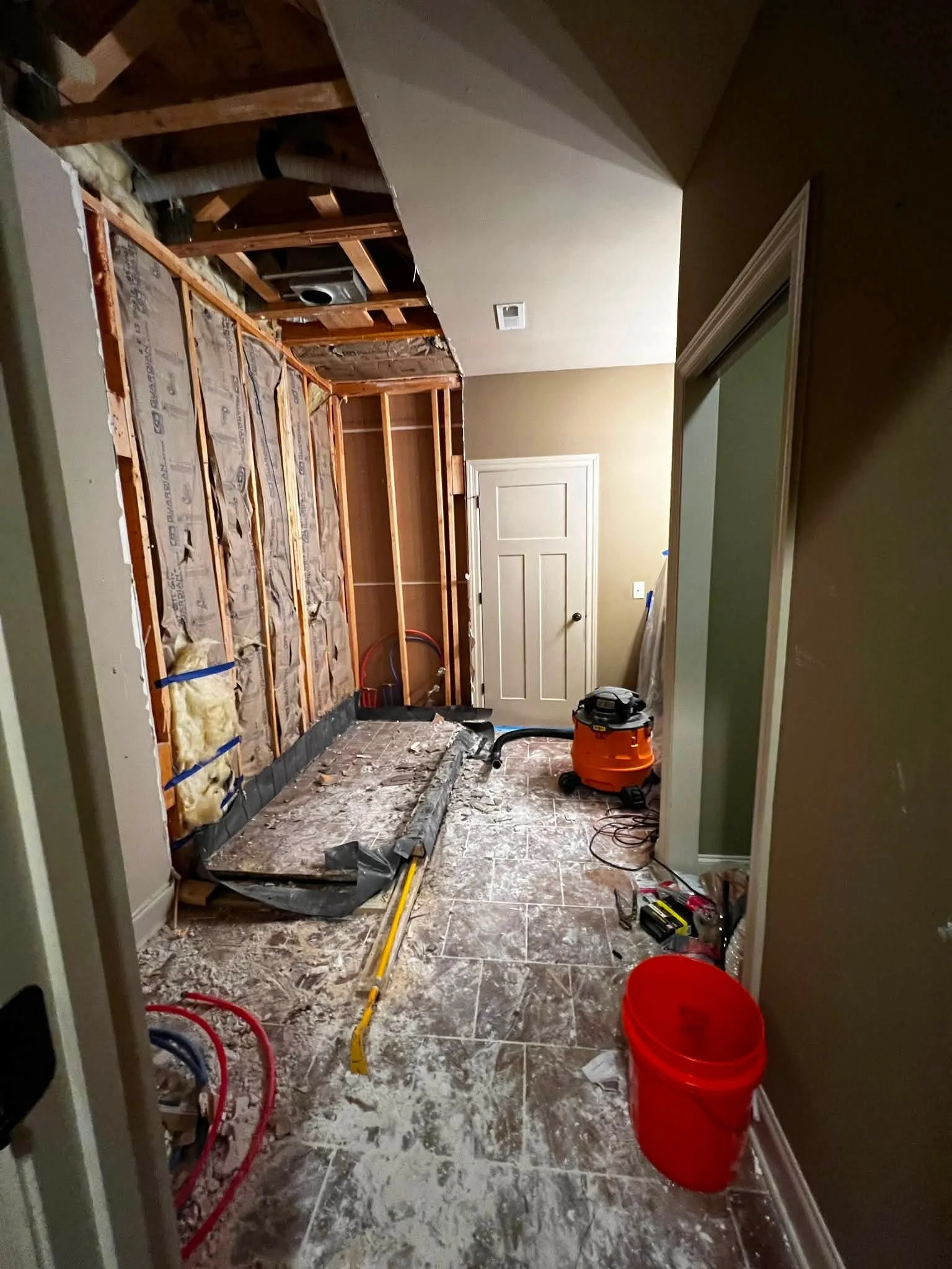 A room under renovation with exposed wooden wall framing, construction tools, and debris on the floor. A cleaning vacuum and a red plastic bucket are visible, along with a closed door in the background.