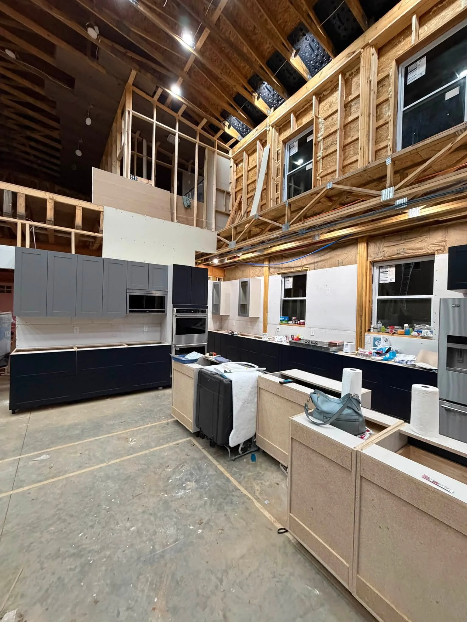 Interior of a building under construction with unfinished walls, exposed wooden framing, and some installed kitchen cabinets in black and grey colors. Construction materials and tools are scattered on the floor.