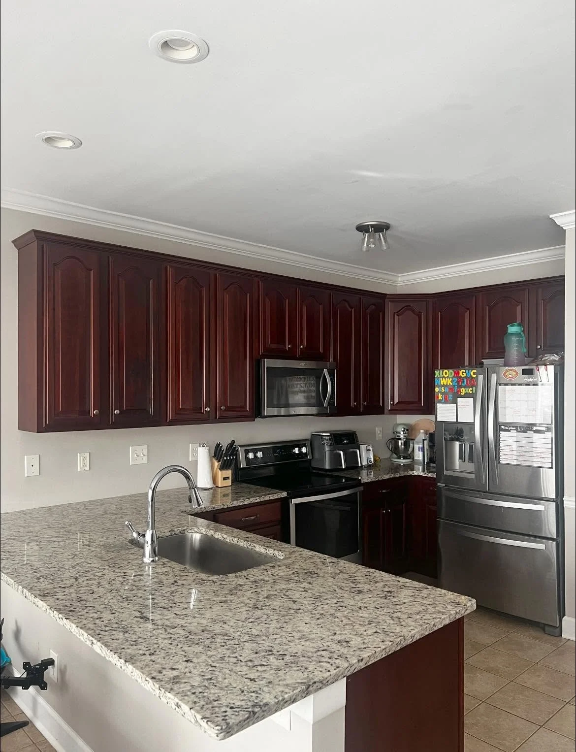 Kitchen with granite countertops, dark wood cabinets, stainless steel appliances including a refrigerator and microwave, a black stove, a toaster, and a stand mixer. There are various kitchen utensils and items on the counter.