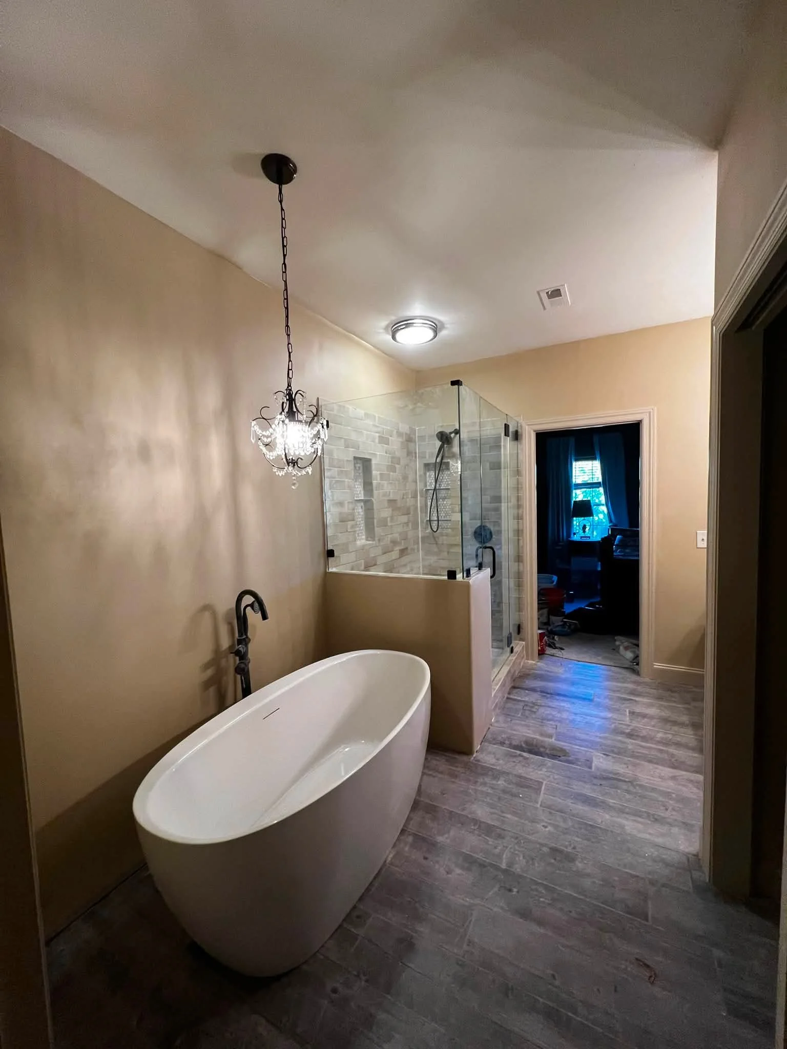 Modern bathroom with a freestanding bathtub, glass shower enclosure with tiled walls, chandelier, and wood-like flooring.