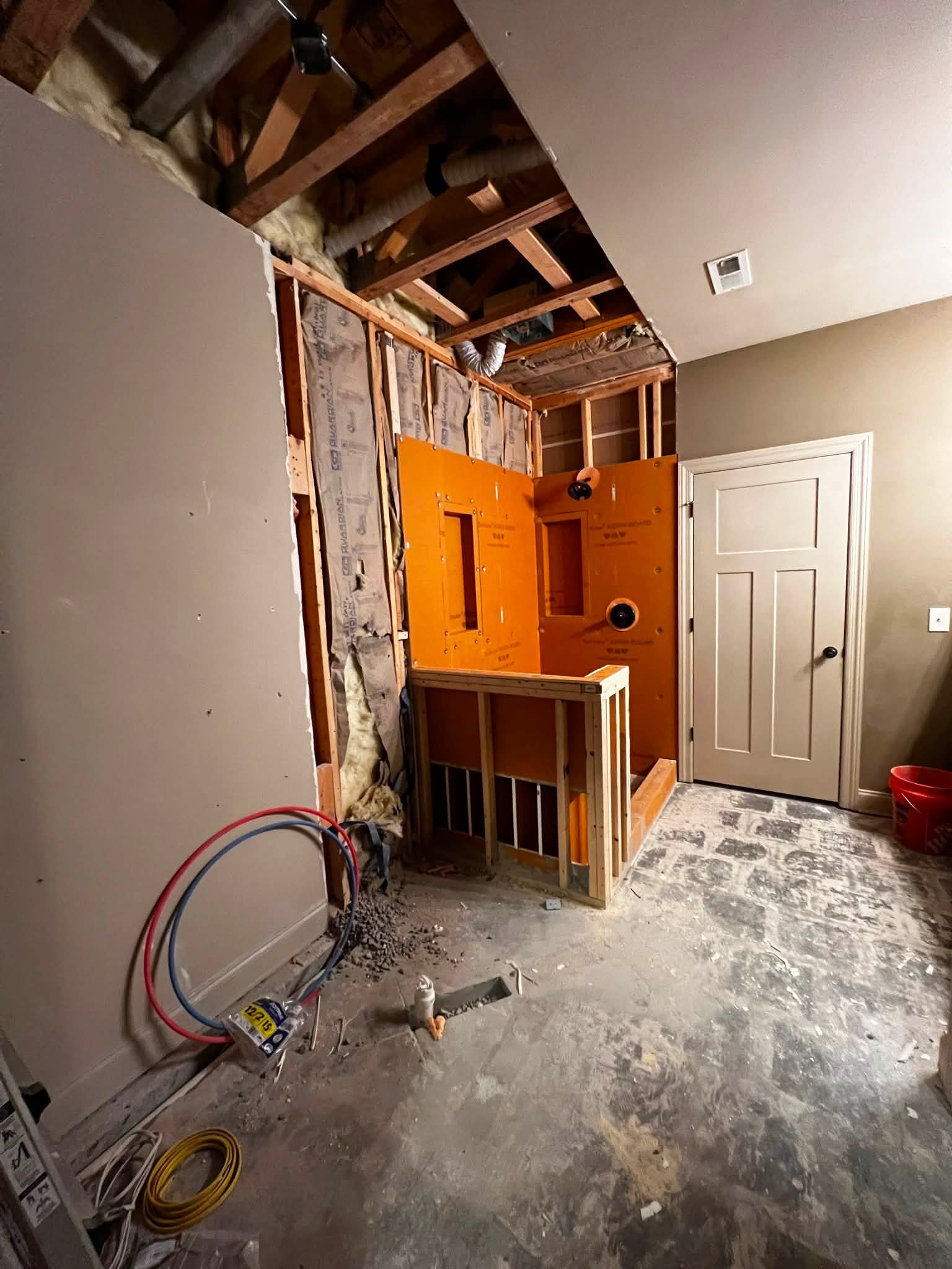Room under renovation with exposed wooden framing, insulation, and electrical wiring visible. The floor is unfinished concrete, and there is a closed door in the background.