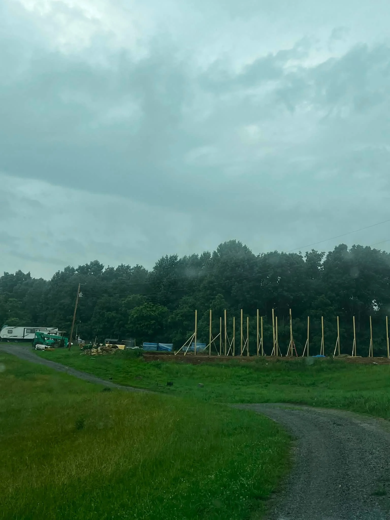 A grassy area with a dirt path, trees in the background, a row of wooden poles, and construction equipment under a cloudy sky.