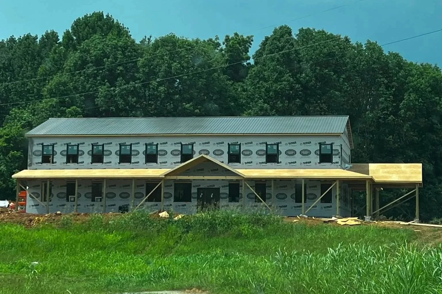 Under construction two-story house with wrapped exterior and a metal roof, surrounded by green grass and trees.