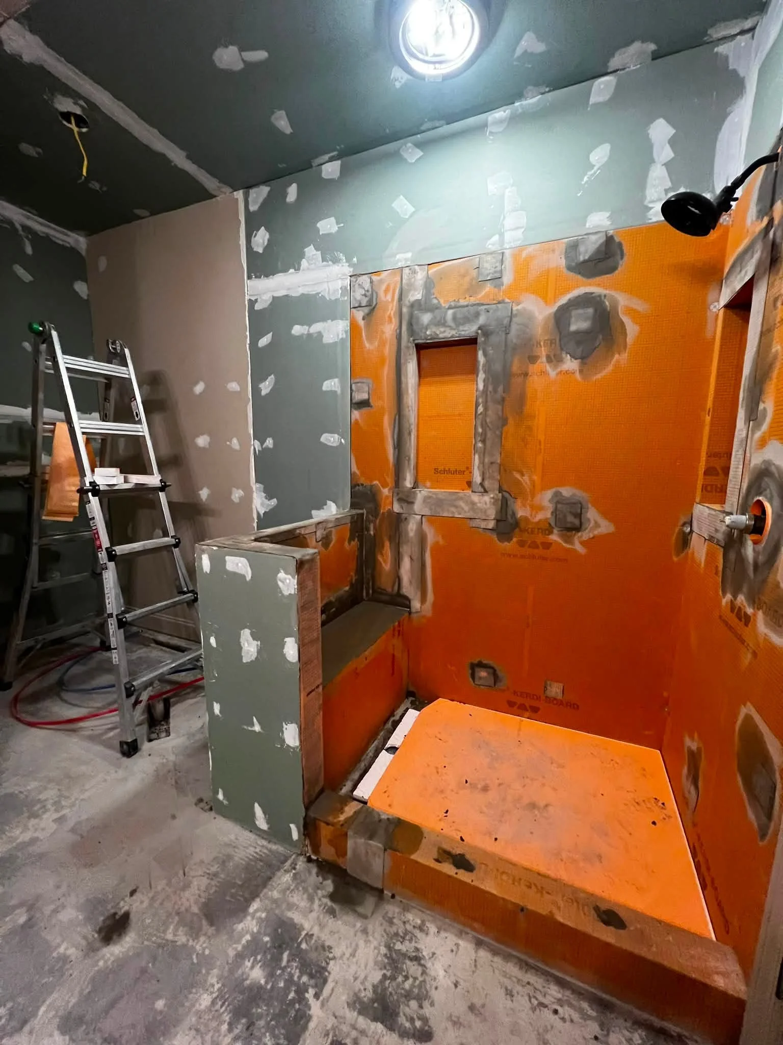 Construction site with drywall installation, orange waterproofing panels, ladder, and unfinished floor.