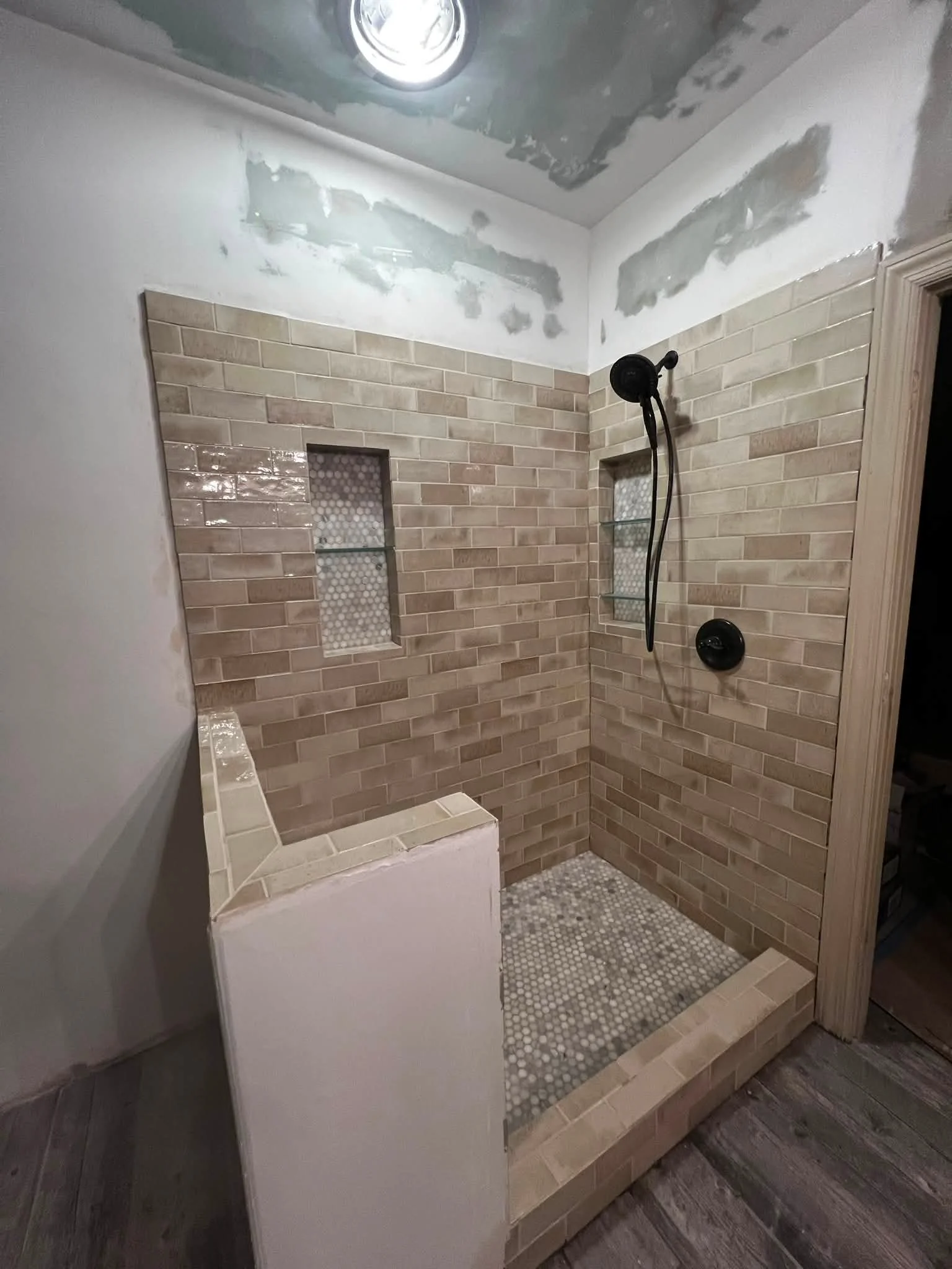 A bathroom shower with beige tile walls, small round mosaic tile floor, built-in shower niches, and a black showerhead and controls. The ceiling is being refinished with drywall compound.