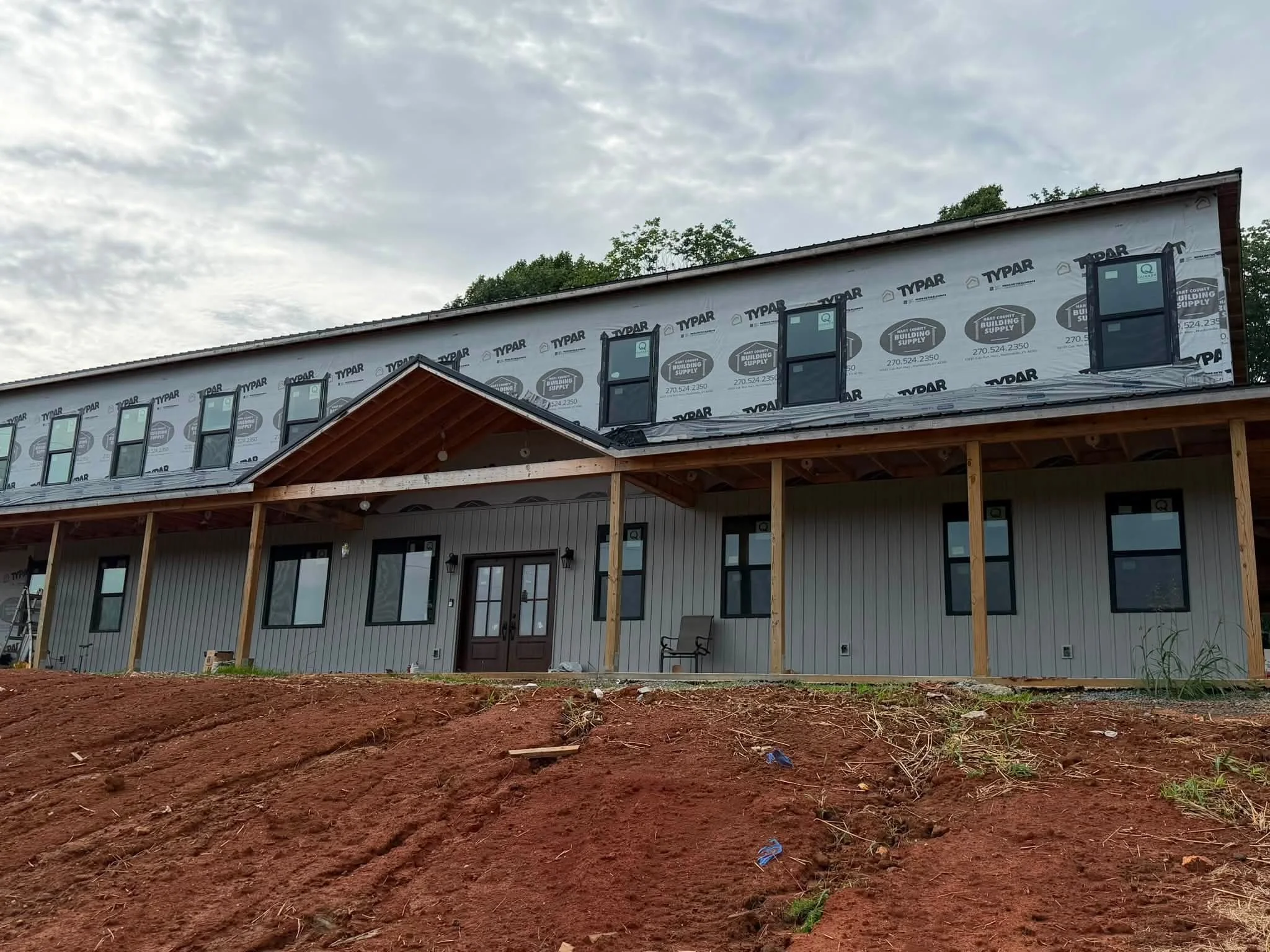 Exterior view of a house under construction with a partially completed roof and multiple windows, with red dirt in the foreground.