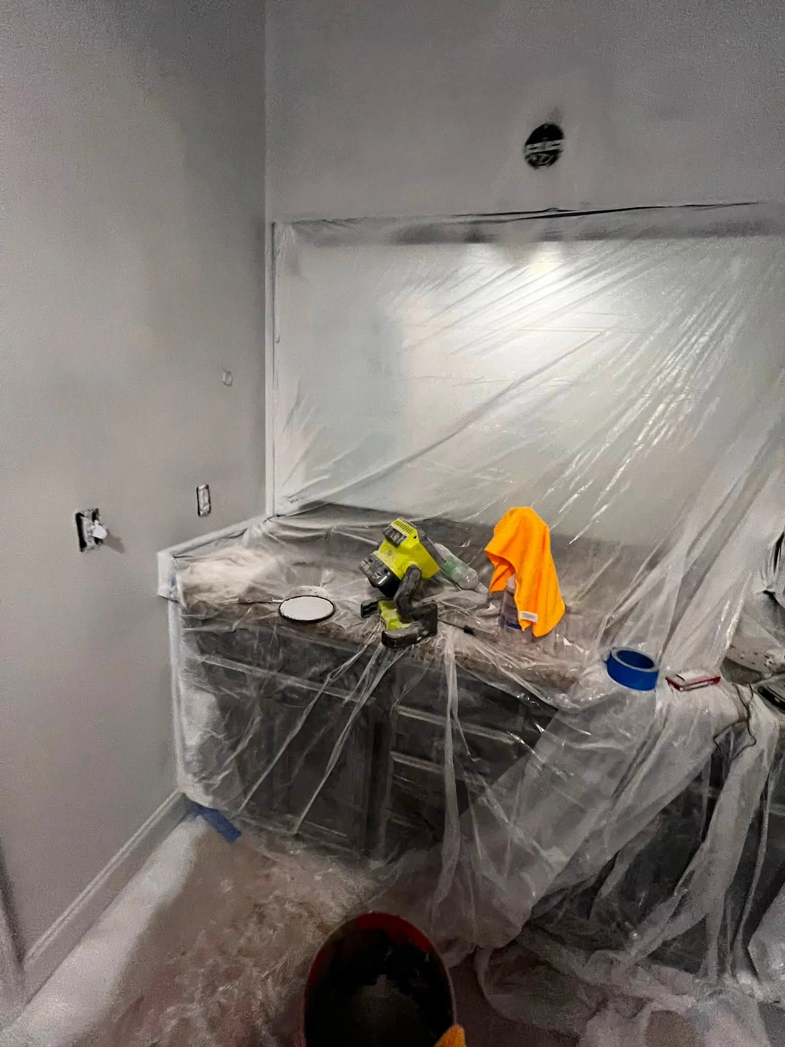 A room under renovation with plastic sheeting covering furniture and floor. Tools such as a drill, a level, and a paint roller are placed on a covered table. An orange cloth and a yellow and black power tool are also visible. The walls appear to be f