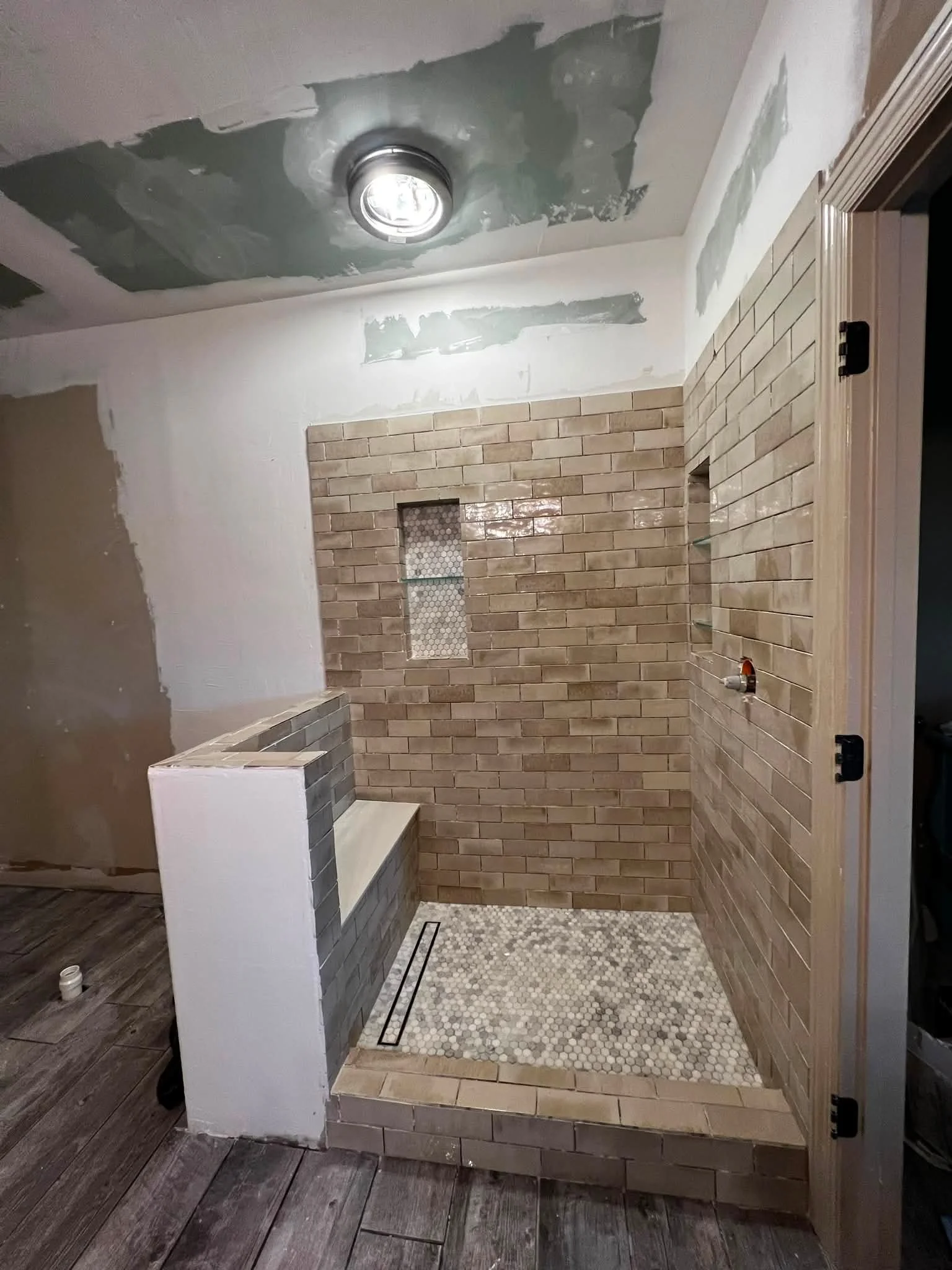 Construction or renovation site of a bathroom shower area with brick wall tiles, a small niche, and a partial view of a wooden floor and wall. The ceiling is unfinished with drywall patches and a recessed light installed.