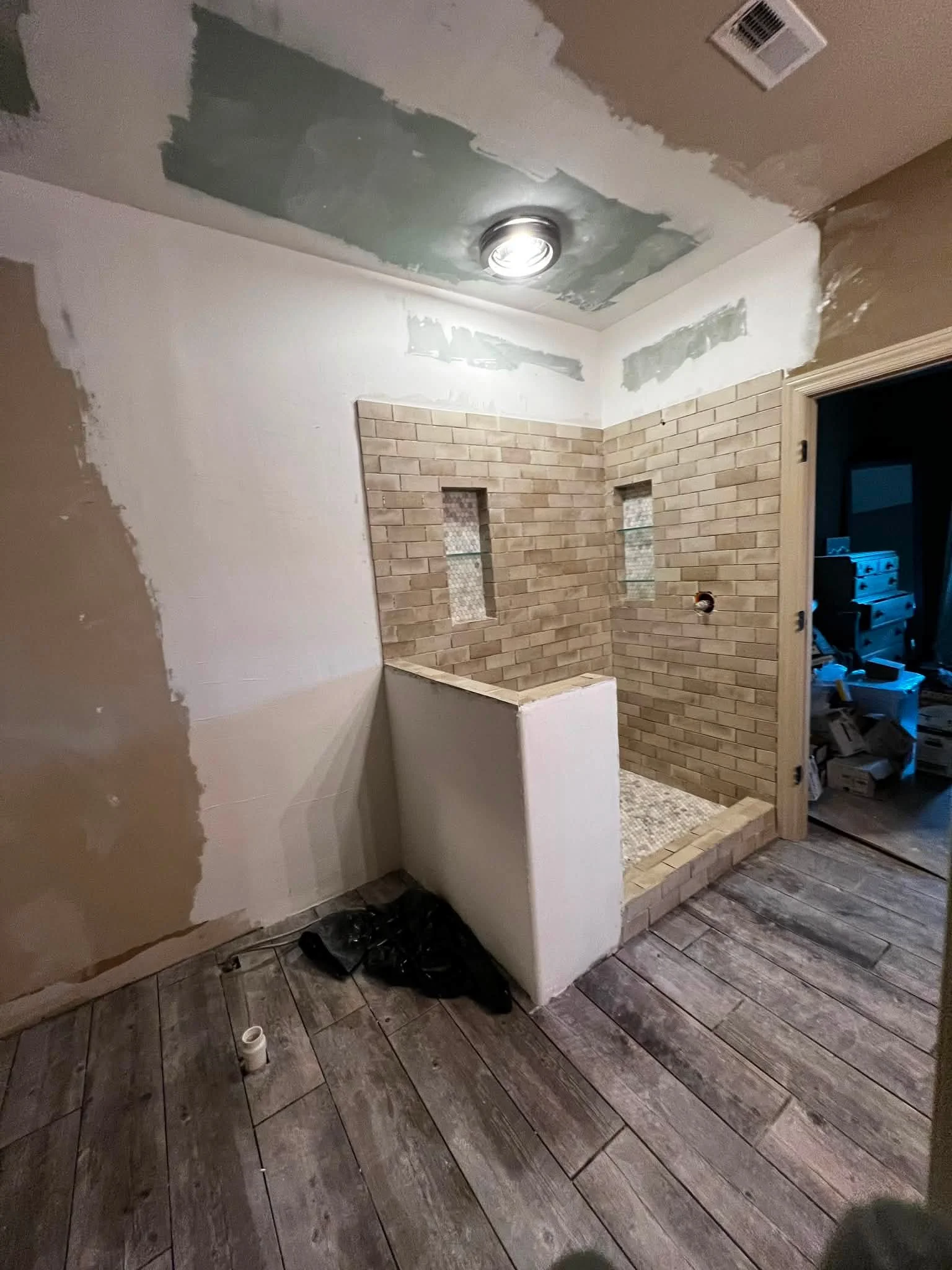 Bathroom under renovation with unfinished ceiling, exposed brick wall, and dark wooden tile flooring. Construction materials and tools are visible.
