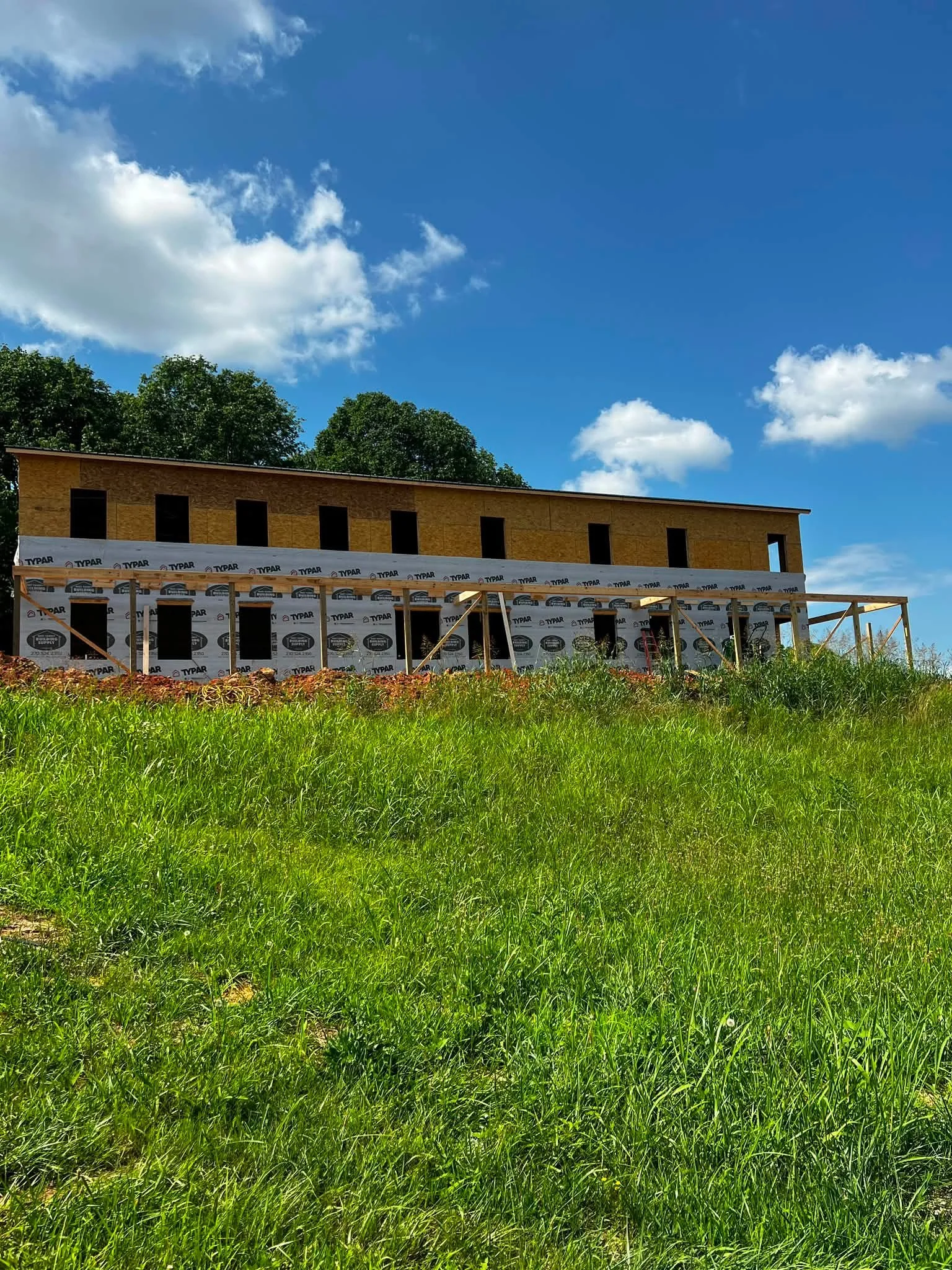 A house under construction with a wooden frame and windbreaker, set against a background of trees and a bright blue sky with scattered clouds.