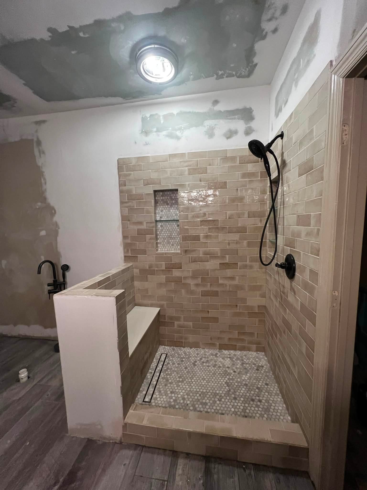 Bathroom shower area under renovation with partially finished tiled walls, a built-in niche, and a framed glass shower door opening, with a black showerhead and fixtures visible.