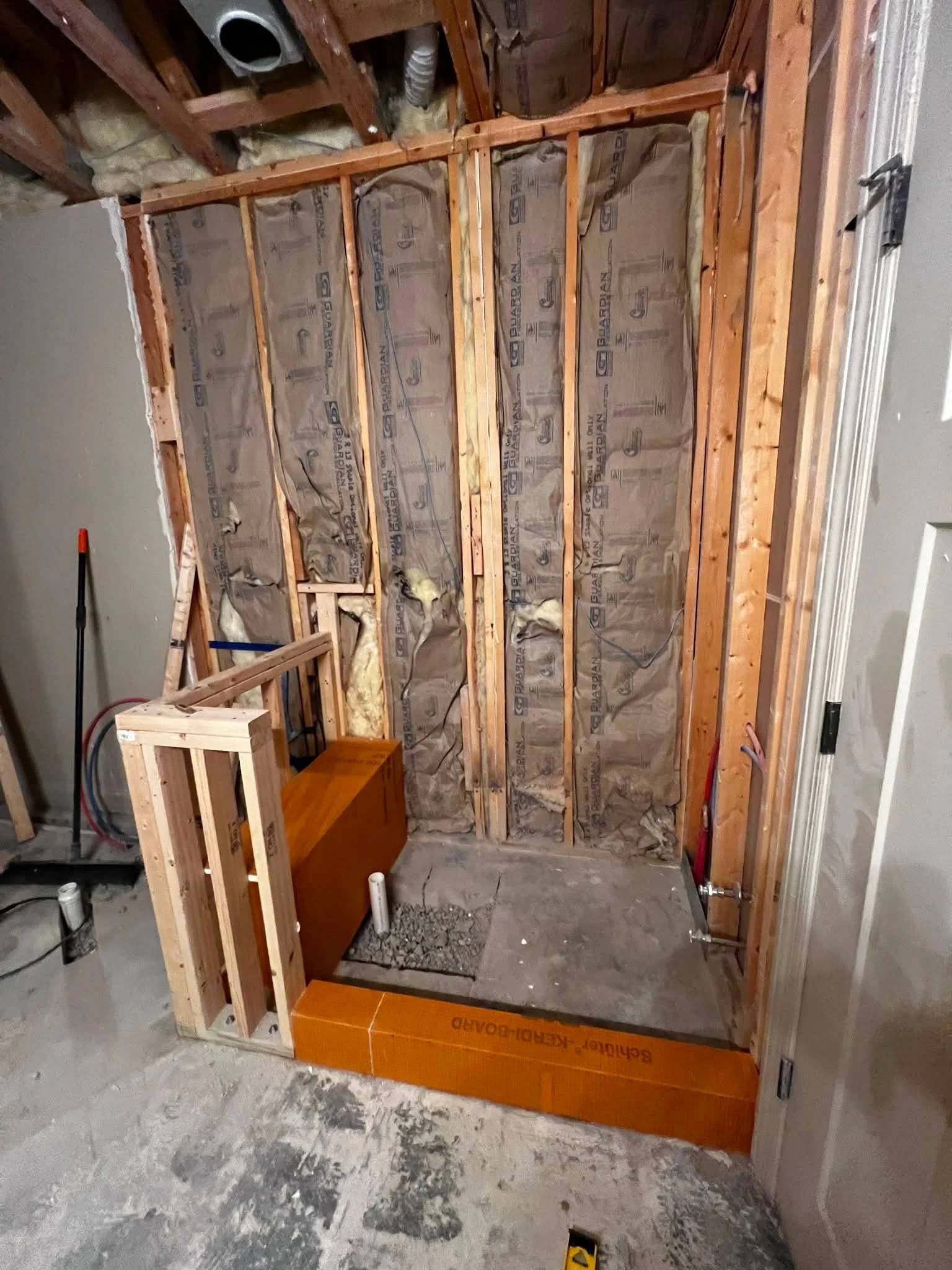 Bathroom under renovation showing exposed wooden wall studs, insulation, and plumbing pipes, with construction materials and tools nearby.