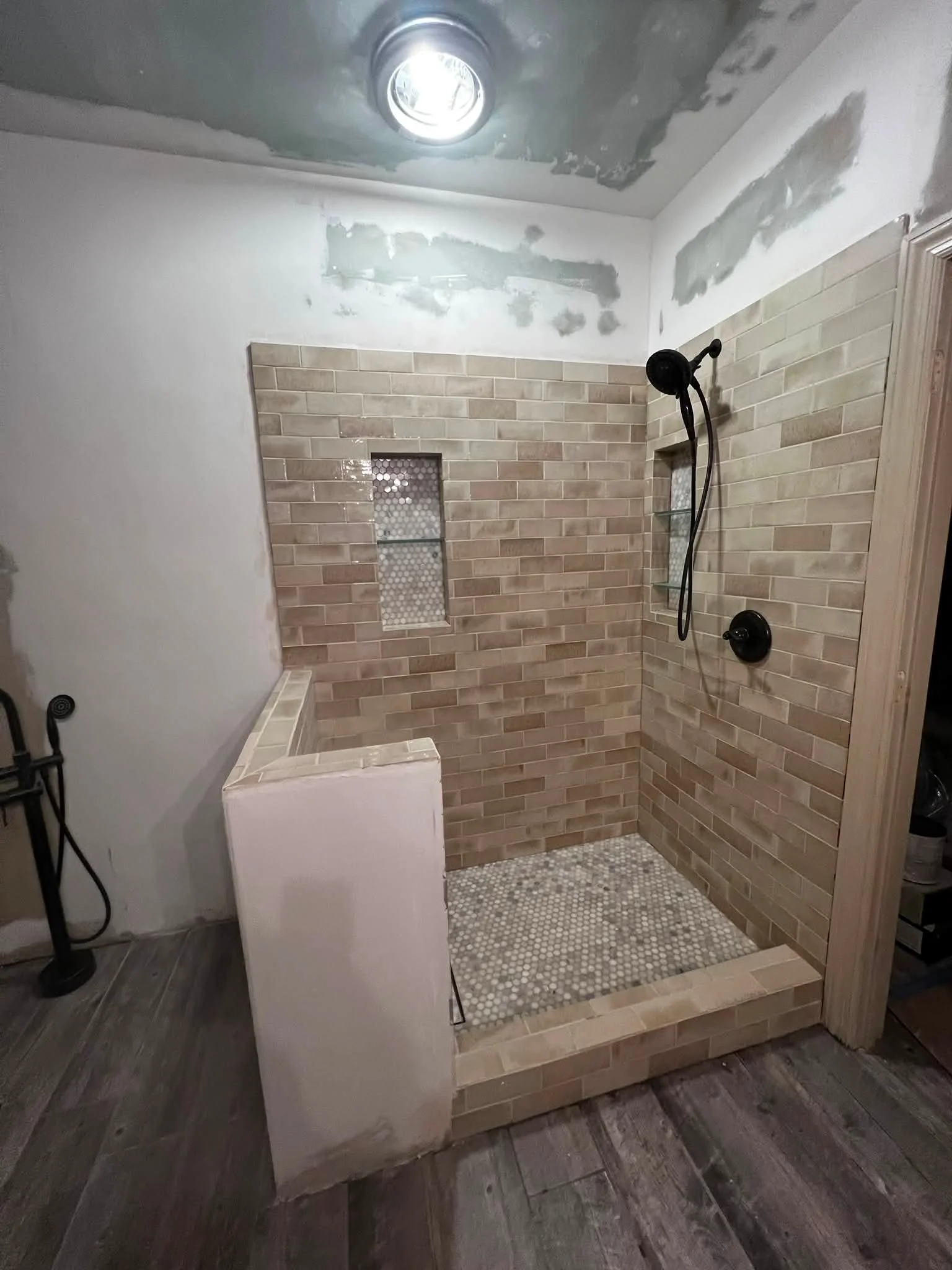 A bathroom with a walk-in shower featuring beige brick-patterned tiles on the walls and small mosaic tiles on the shower floor. There is a small built-in niche with shelves inside the tiled wall. The ceiling appears to be unfinished with patches of d