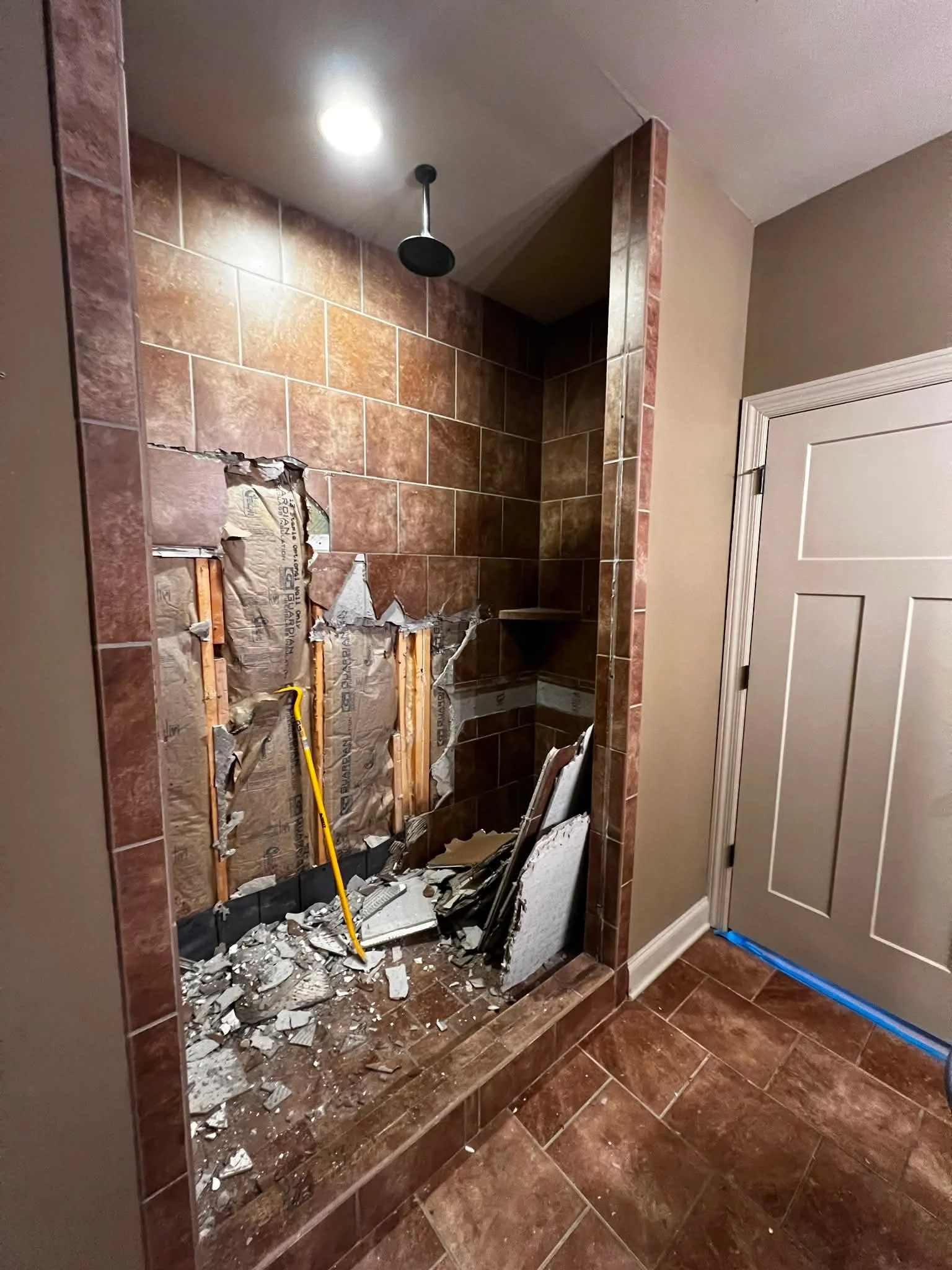 Bathroom with section of tiled wall torn open, rubble on the floor, a yellow crowbar, and exposed wall studs, indicating construction or repair work.