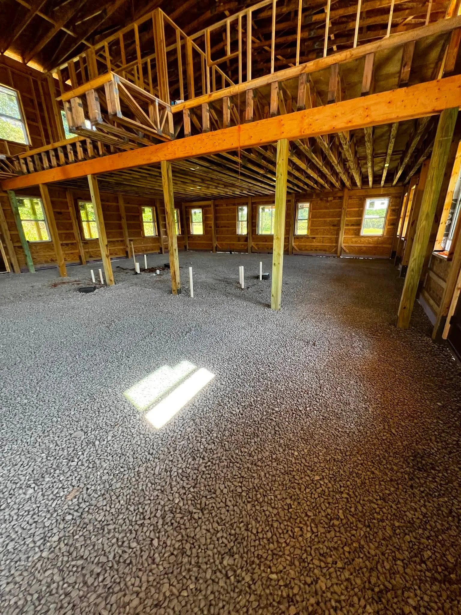 Interior of a house under construction with exposed wooden framing, multiple windows, and a gravel floor.