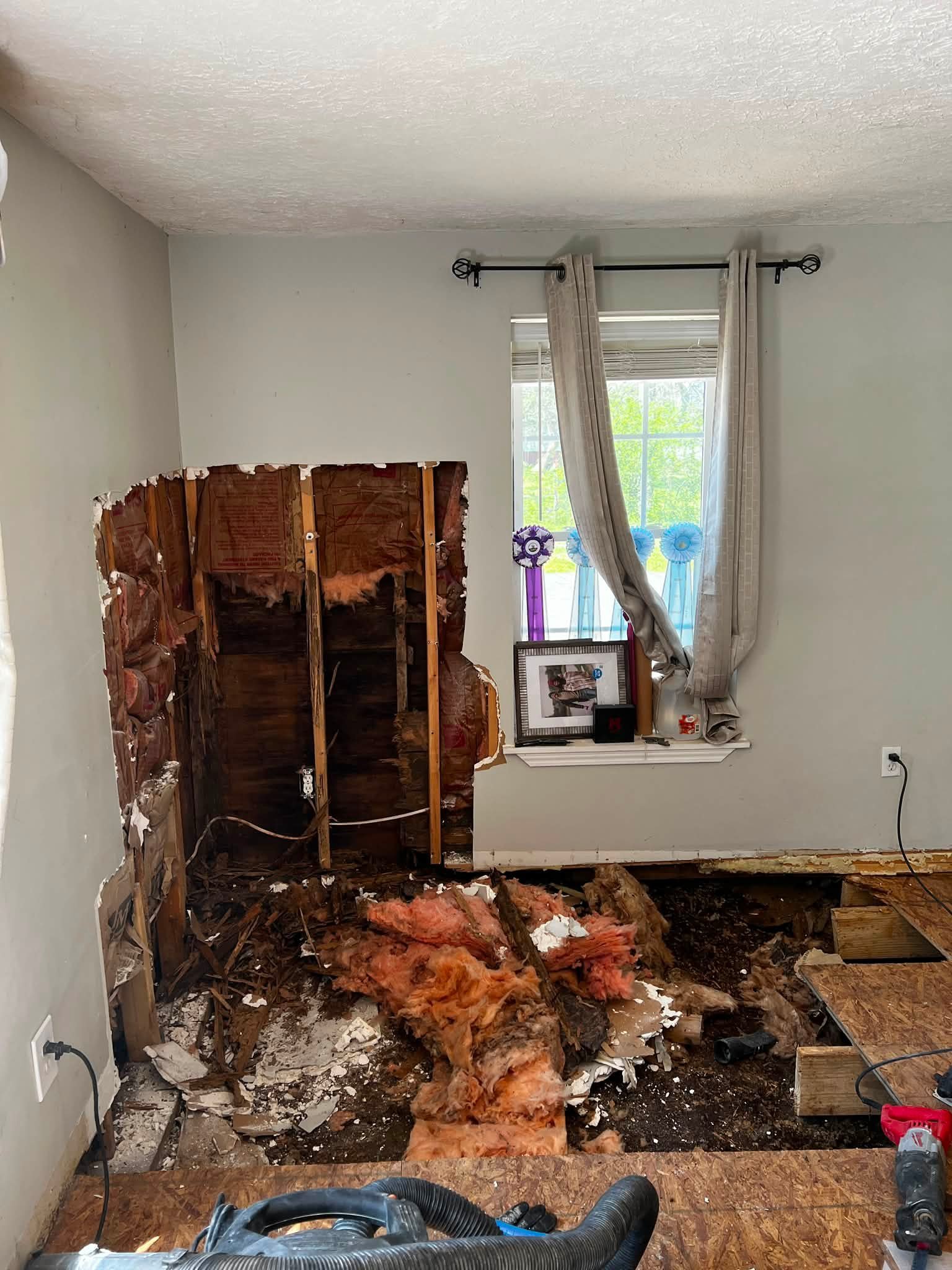 Room with a section of wall removed, exposing wood framing and insulation. The floor is covered with debris, insulation, and damaged drywall. There is a window with curtains and ribbons on it, and tools are scattered around the floor.