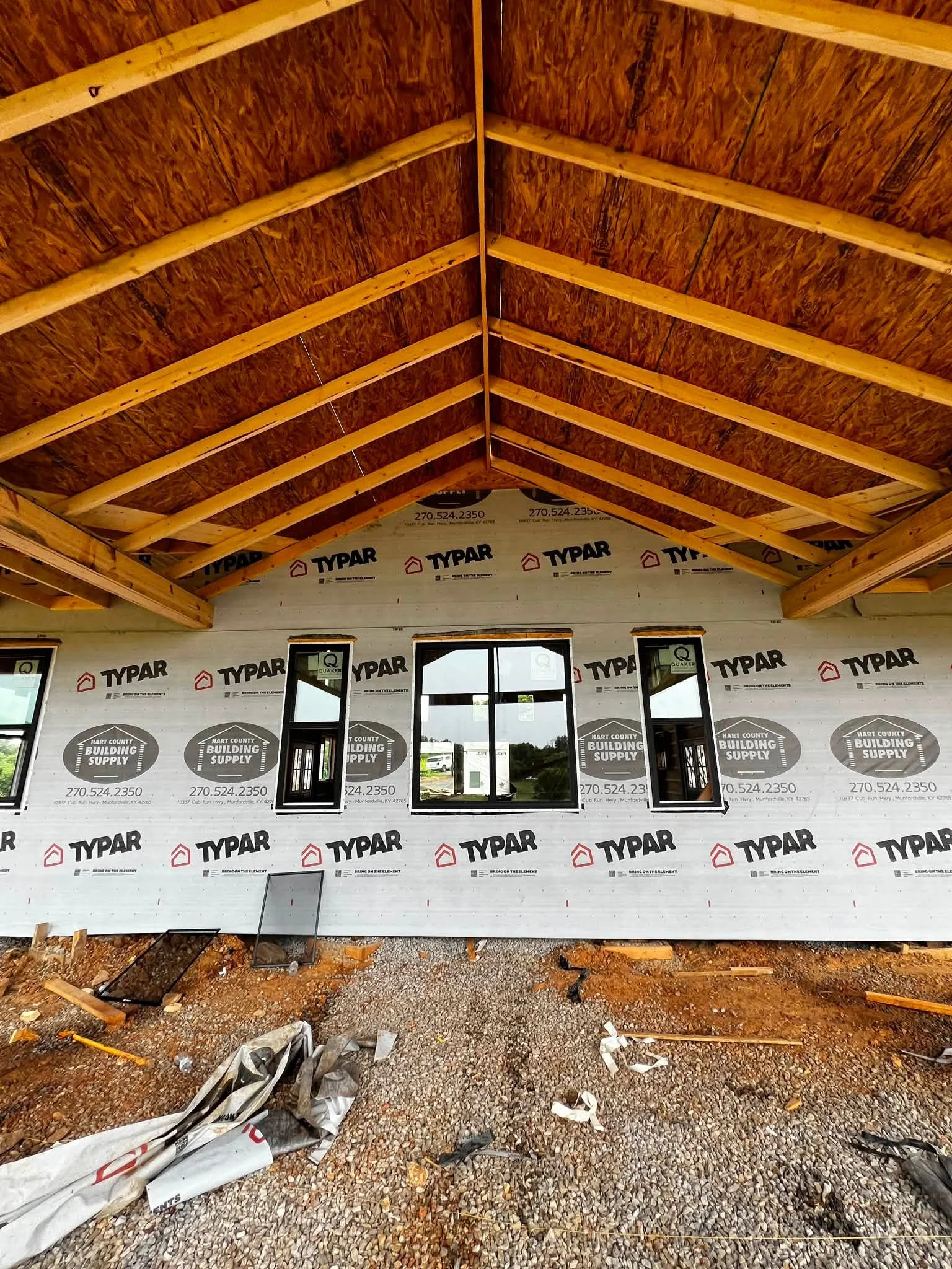Interior of a house under construction showing a second-story ceiling with exposed wooden beams and plywood sheathing. The lower part of the wall is wrapped with Typar building wrap, with three rectangular windows installed. Construction debris is sc