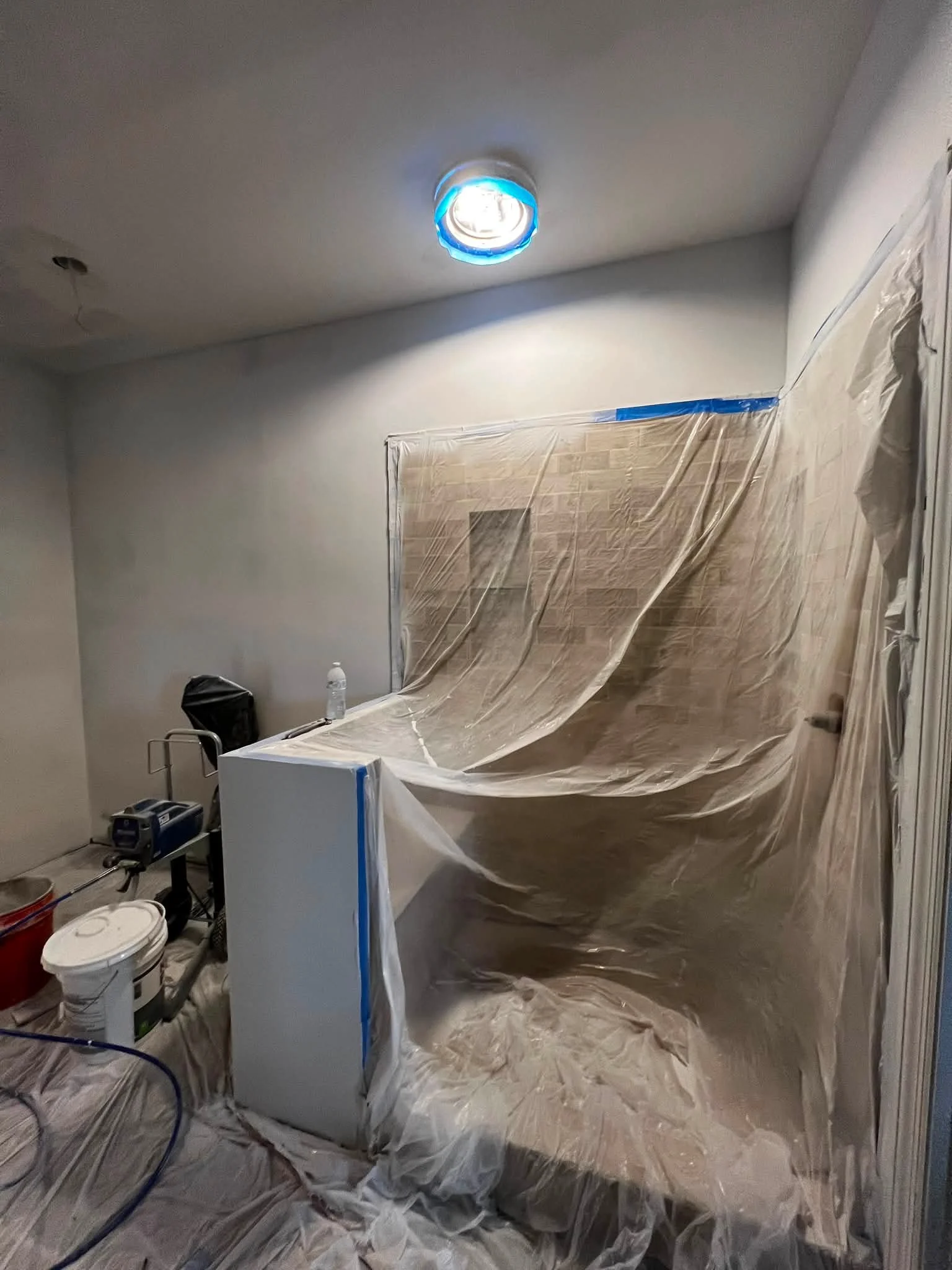 Room under renovation with plastic sheeting covering the floor and wall. A recessed ceiling light is installed, and tools and supplies are on the floor, including a bucket, a small white refrigerator, and a water bottle.