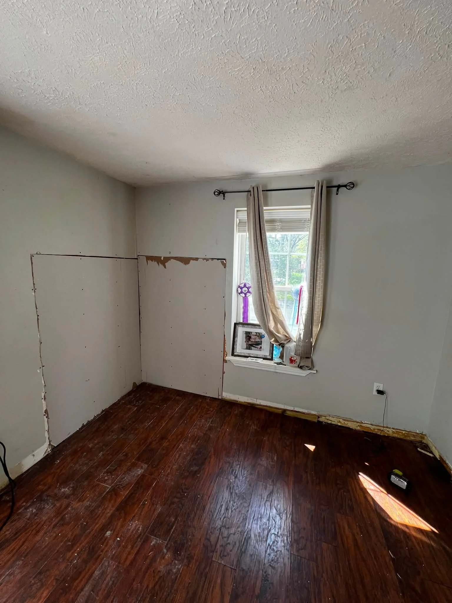 A room with dark wood flooring, a window with partially drawn beige curtains, and a wall with a large, partially removed section of drywall. The room appears to be under renovation or repair.