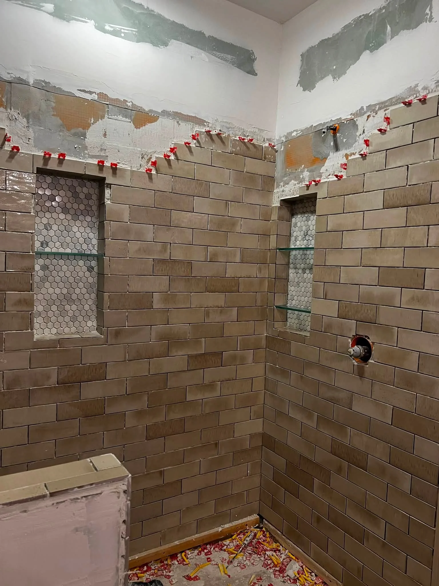 A bathroom under renovation with brown subway tiles on the walls and unfinished areas on the upper walls and ceiling. Red clips hold backsplash tiles in place while grout or mortar dries, and there are construction materials and debris on the floor.