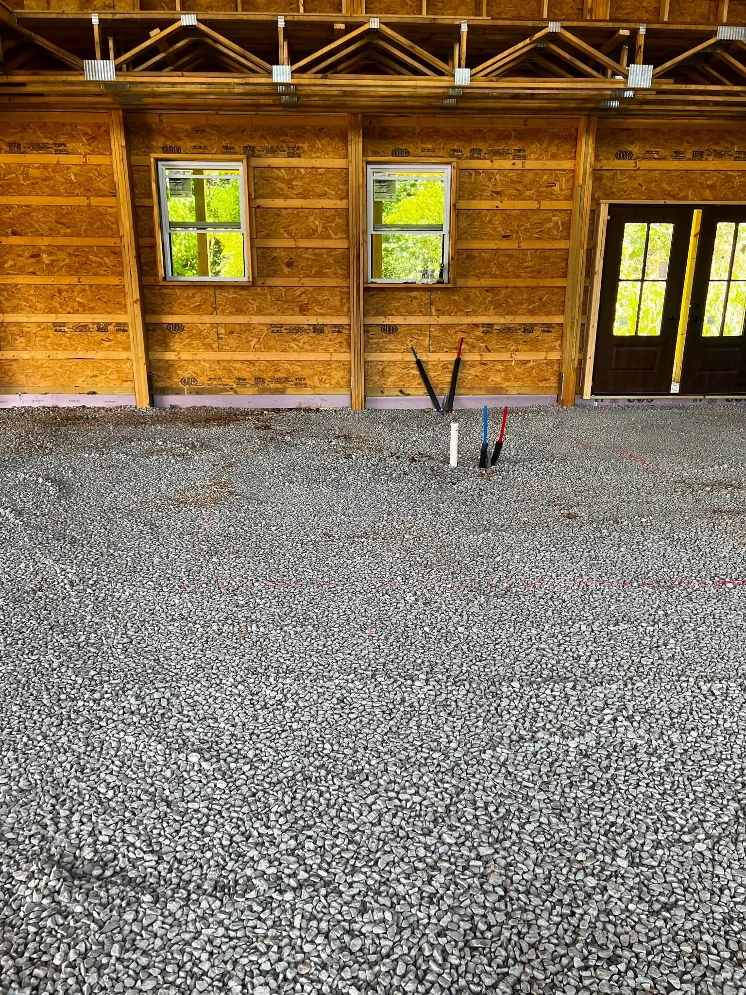 Interior of a house under construction with exposed wooden framing, two small windows, a door with glass panes, and a gravel floor.