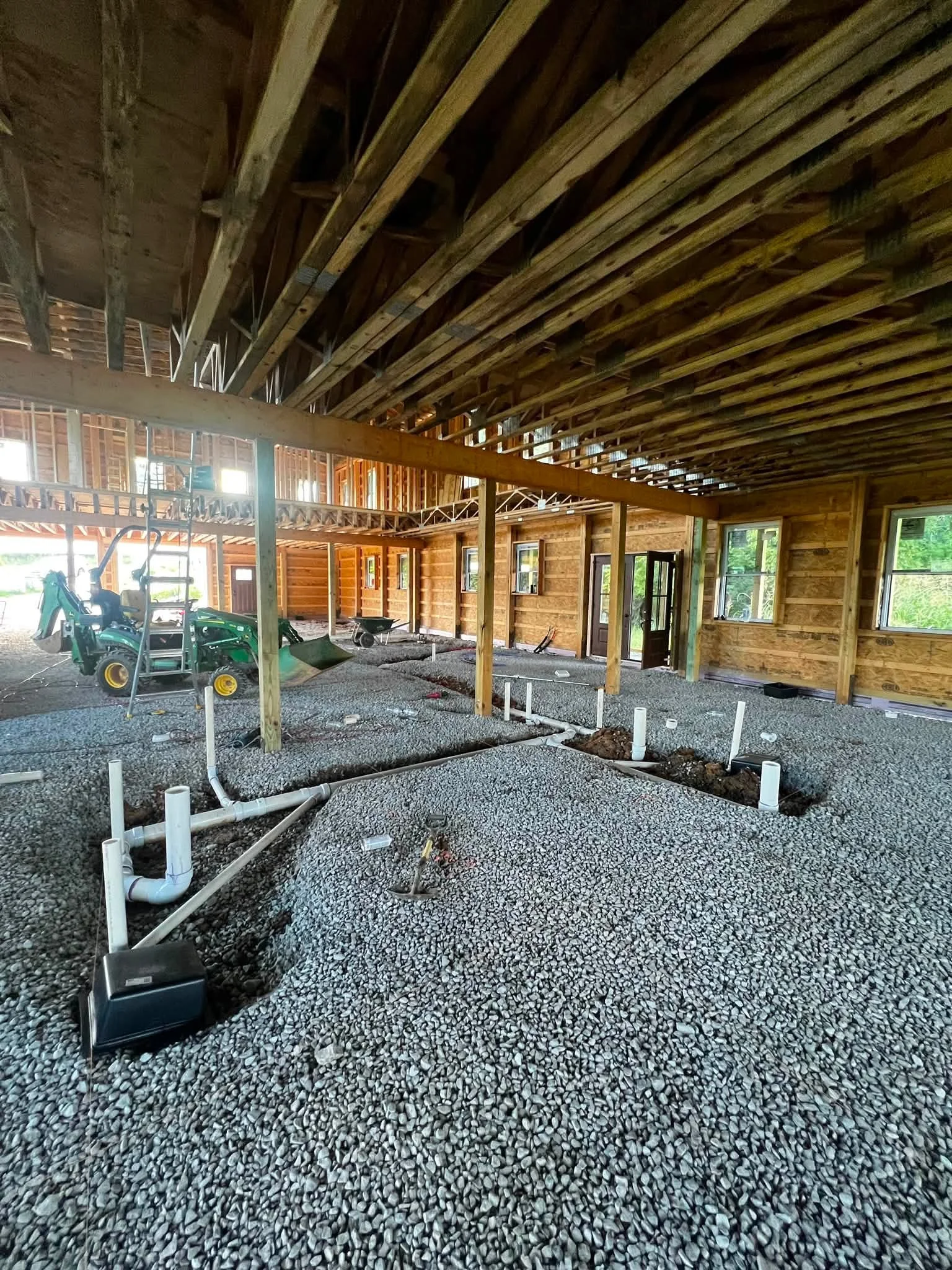 Open construction site with exposed wooden beams, metal framing, and gravel floor with plumbing pipes and machinery.